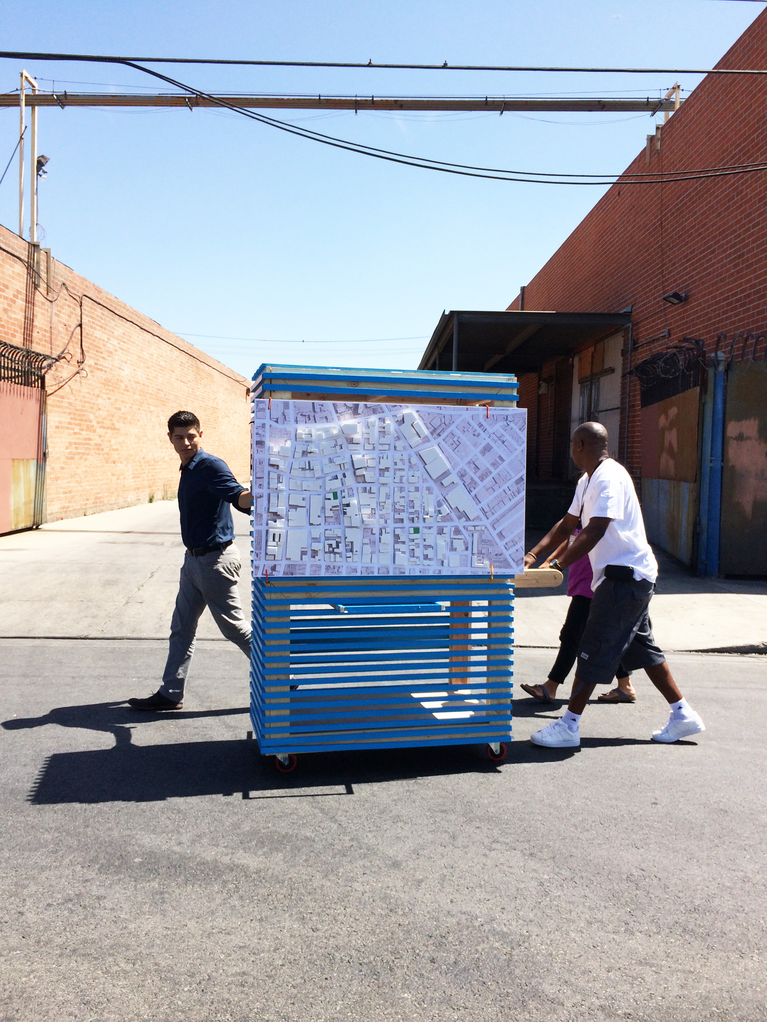 Photo shows Anthony, Saul, and Leslie pushing a blue painted handmade cart with a big model of the Skid Row neighborhood mounted it through the streets of Skid Row to engaged people on the street.