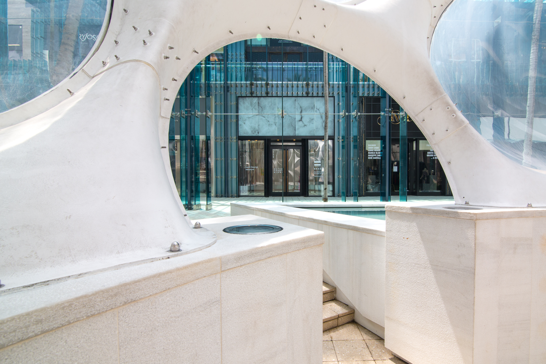 Nadir Sourgi, Installation view of Metonym at Bas Fisher Invitational as part of Waterproof Miami, Photo by Monica McGivern (2022). Peering from half of an oval shaped entrance, the facade of a gallery that is primarily glass is revealed in the distance.
