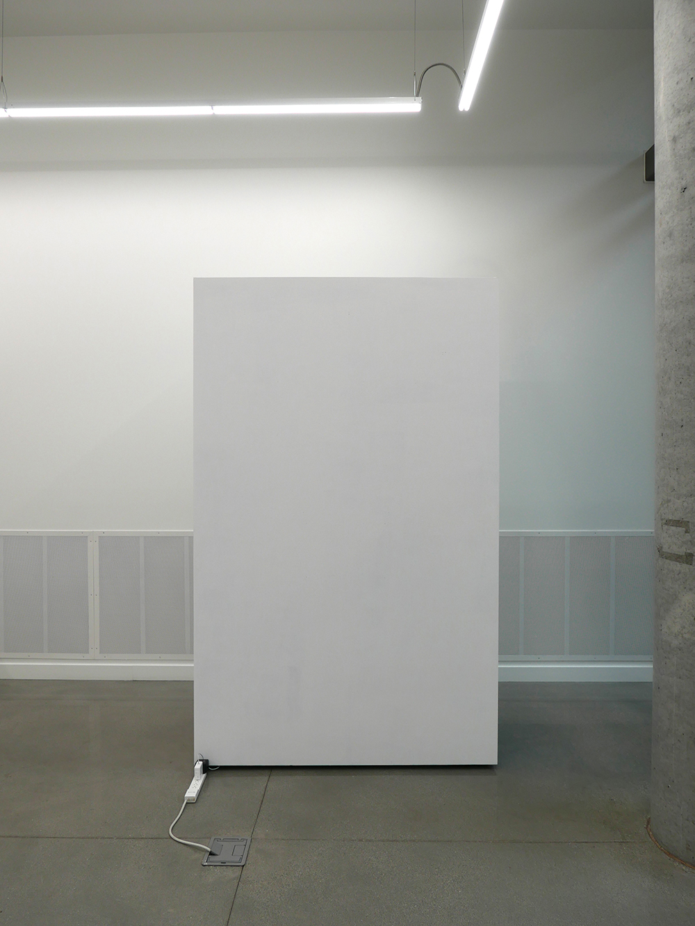 View of a blank wall in a gallery. Electric cables and chargers extend from beneath the wall and appear to be plugged into the power outlet.