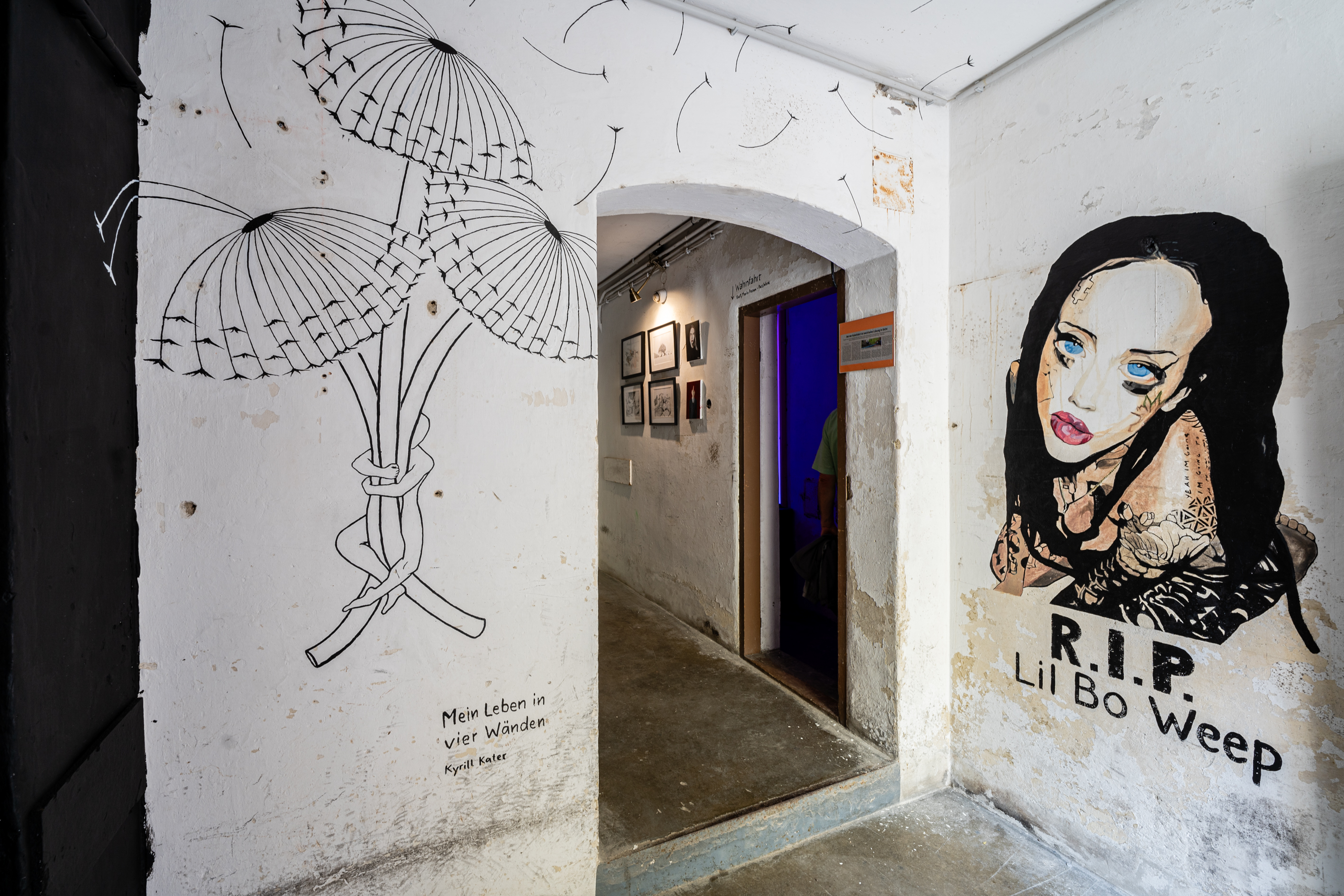 Bild 1 zu Mein Leben in vier W&auml;nden: Vorraum des Hauses Reality 1: An der rechten Wand eine Malerei einer Frau mit schwarzen langen Haaren. Darunter steht R.I.P. Lil Bo Weep. An der Wand gegen&uuml;ber der Eingangst&uuml;r sind drei verbl&uuml;hte L&ouml;wenzahnbl&uuml;ten mit schwarzem Pinselstrich gemalt, die von einer vereinfachten Person am Stiel zusammengehalten werden. Rechts der T&uuml;rbogen, der in das Treppenhaus des Hauses f&uuml;hrt. 