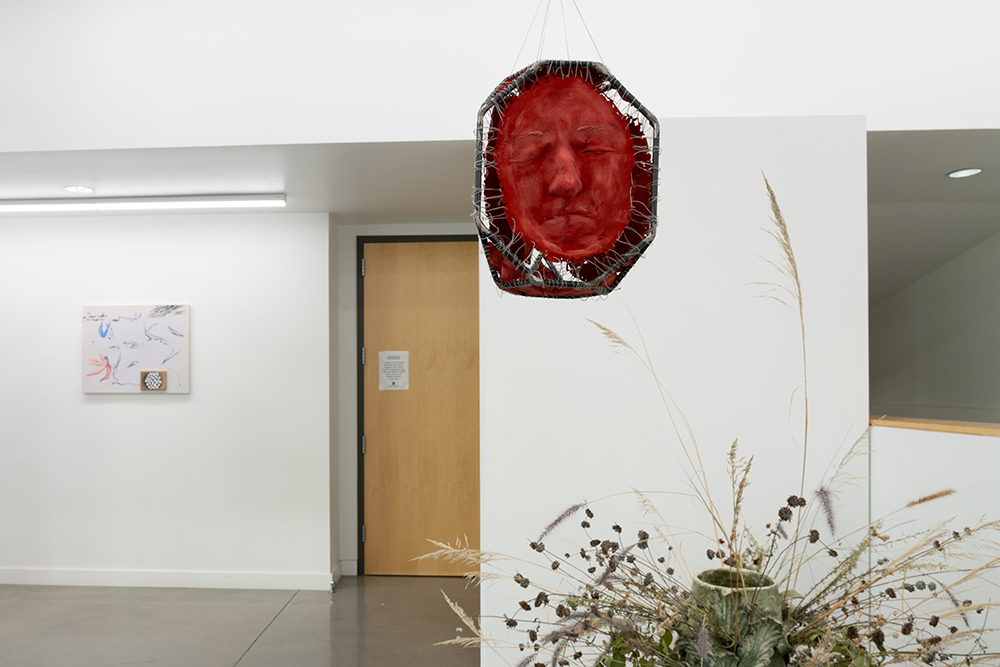 A painting hanging on the wall in distance, a hanging sculpture of a frown face in a metal frame, and a partial view of a vase with dry plants and flowers.