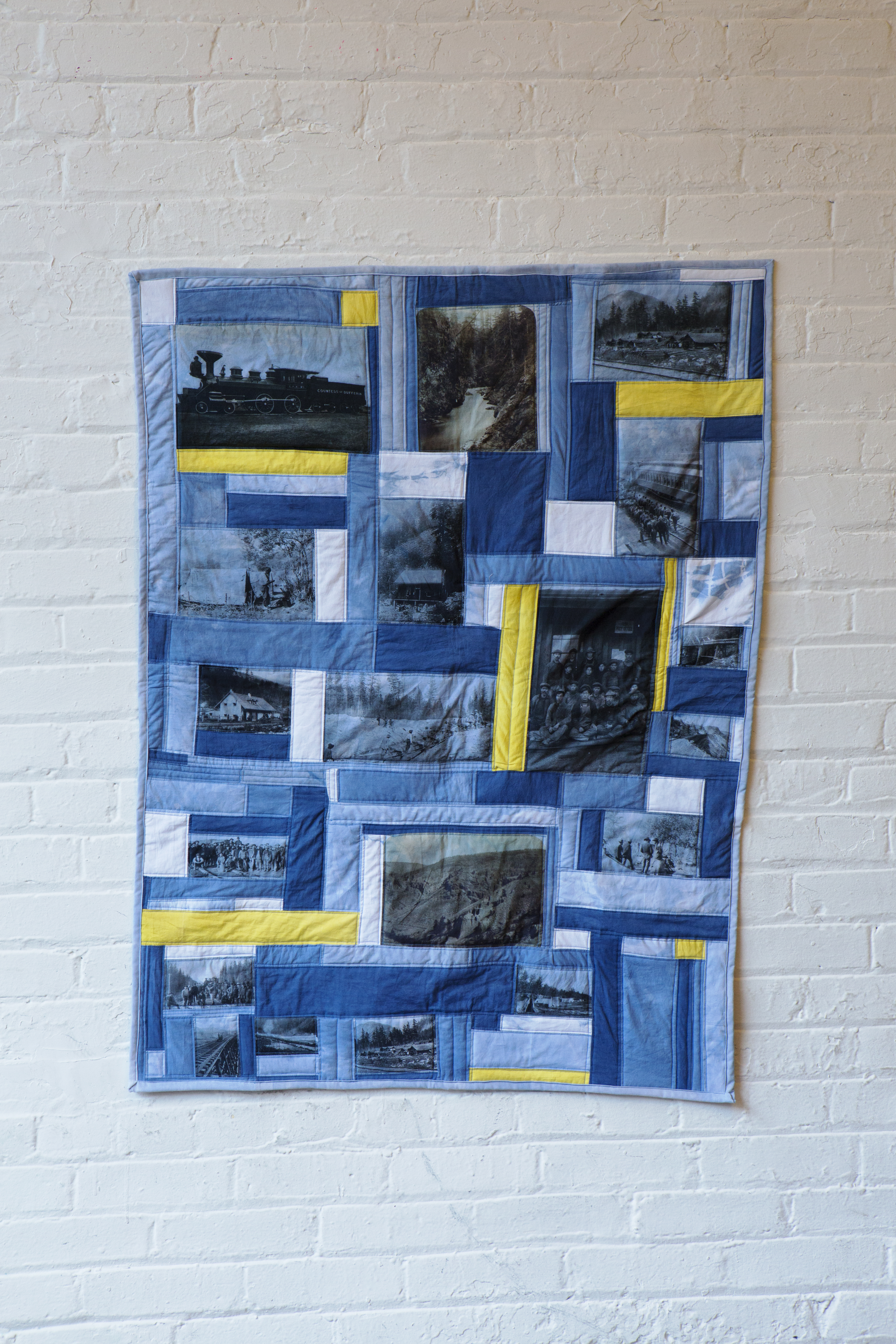 A quilt of dark blue, light blue, tie dye blue and yellow panels. On several panels are black and white images of scenes from the building of the Canadian Pacific railway, including the wilderness landscapes, a steam engine, cabins, and group photos of the Chinese Canadian workers.