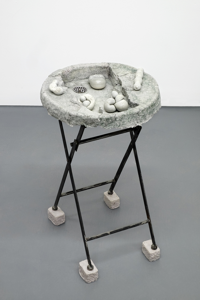 Black folding steel legs stand atop rectangular gray paper pulp bricks. A circular gray top holds 5 smooth rounded concrete forms.