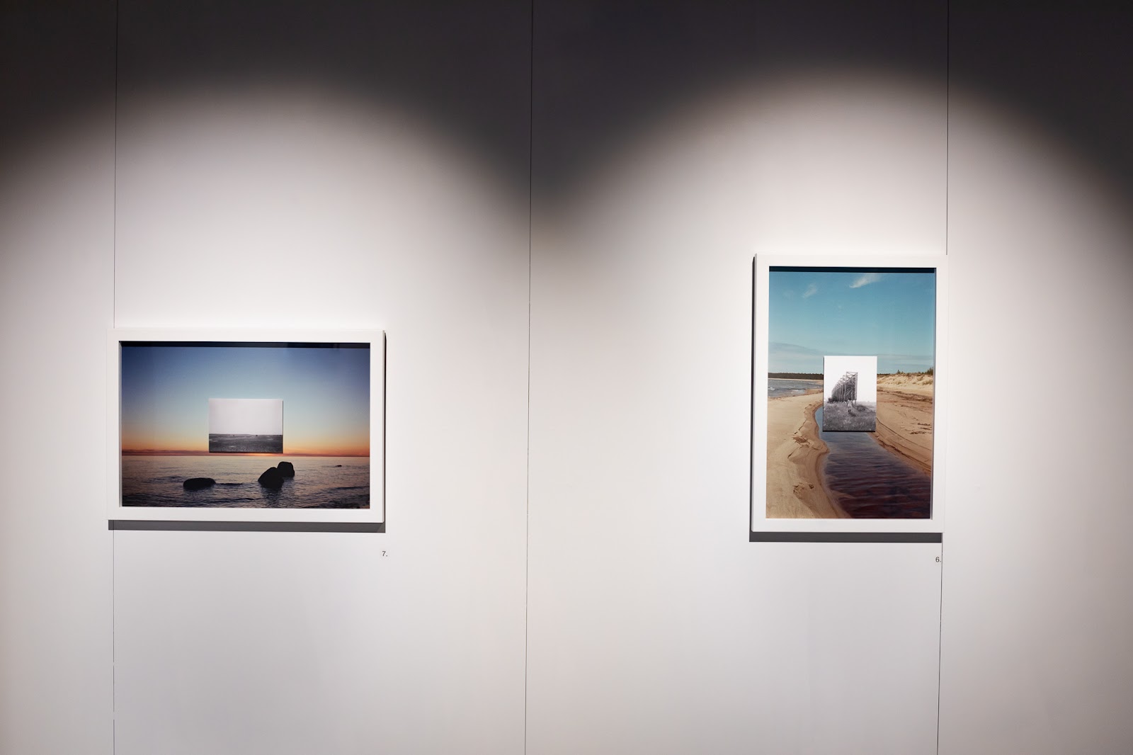 A white wall of a dimly lit gallery space with two framed photographic artworks, the left being oriented horizontally and the one on the right oriented vertically. They both depict empty feeling nature landscapes with smaller images superimposed in their centers. Bright spotlights individually illuminate the area around each work but leave the rest of the space dark.