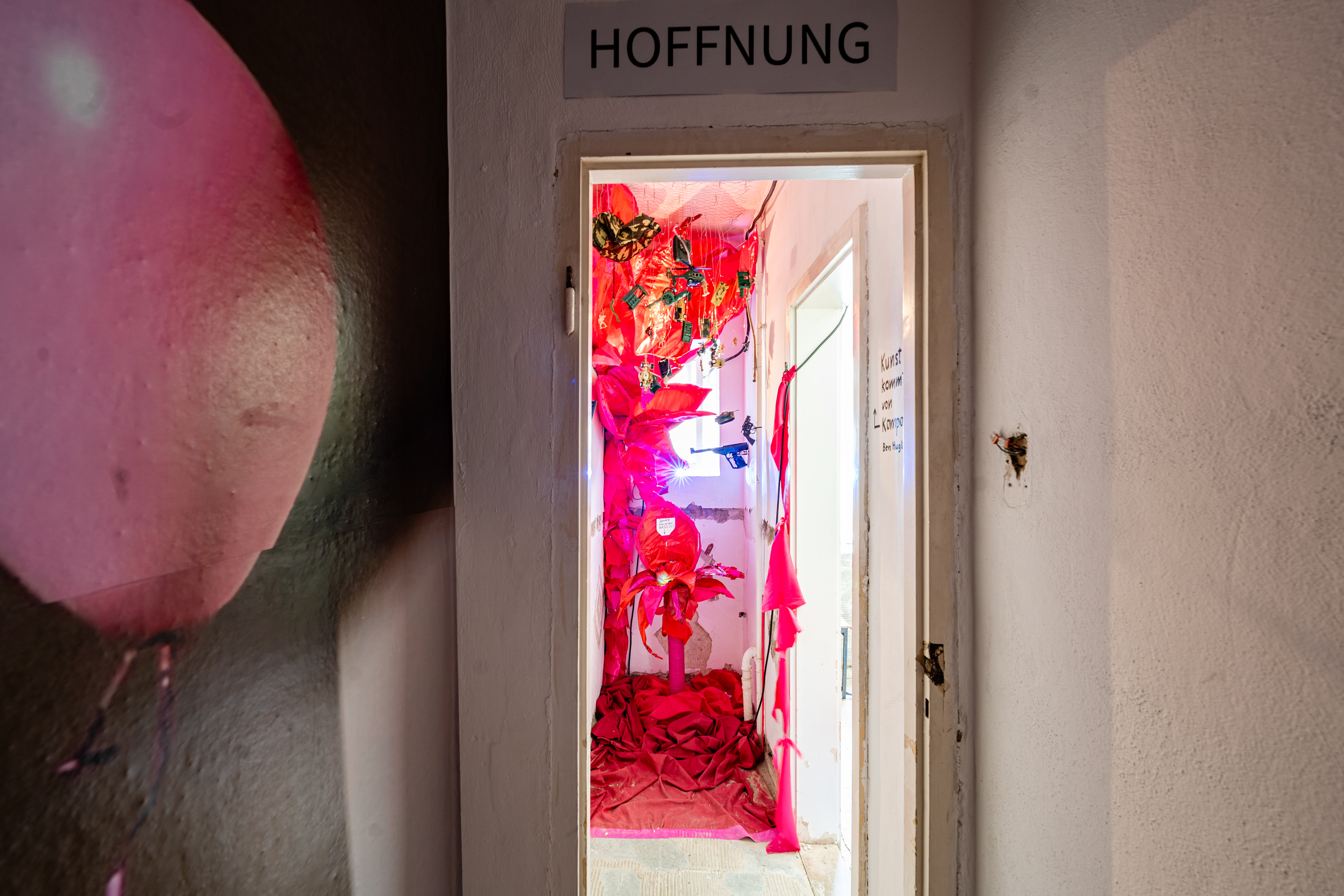 Bild 1 Krieg fressende Pflanze: Blick durch wei&szlig;en T&uuml;rrahmen in schmalen Raum, in dem sich rechts eine weitere T&uuml;r&ouml;ffnung befindet. &Uuml;ber dem T&uuml;rrahmen des Raumes steht in Druckbuchstaben Hoffung. An der linken Wand des Raumes sind aus rotem Stoff  gro&szlig;e ovale Bl&auml;tter einer Pflanze immitiert, die bis zur Decke rankt.  Am Boden befindet sich ebenfalls zerkn&uuml;llter roter Stoff. Von der Decke h&auml;ngen an d&uuml;nnen F&auml;den kleine Soldatenfiguren,  und kleine Waffen, Panzer und Helikopter aus Plastik.