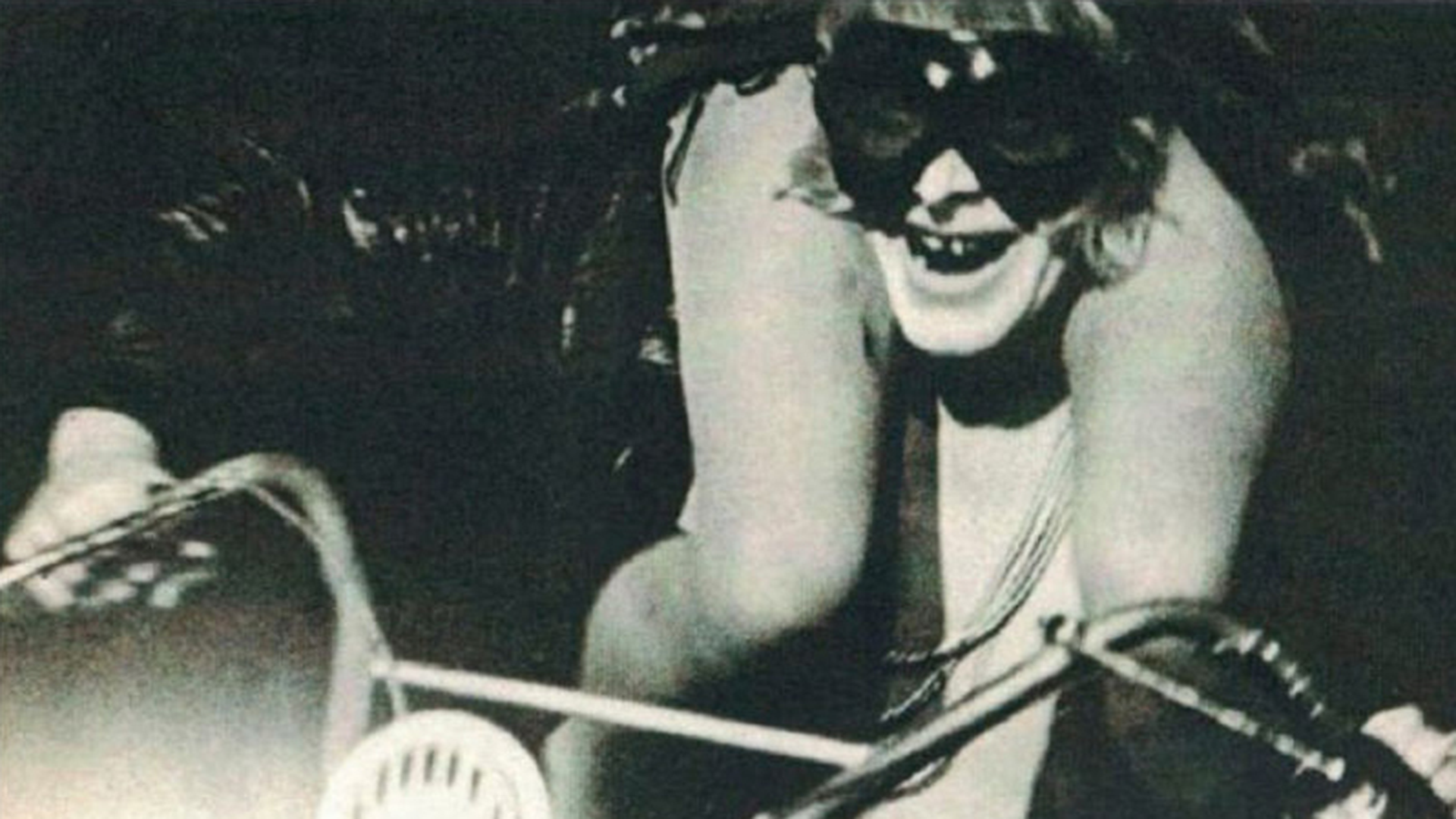 ID: A black and white photograph of a man wearing riding goggles whilst riding a push-bike. The man is smiling, and the bare legs of a woman are visible, she sits on his shoulders and is cropped out of the frame.