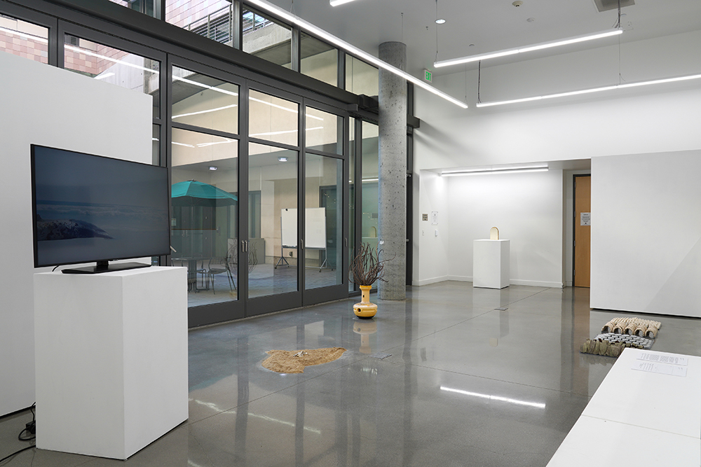 Five artworks are installed in a gallery: one on a TV screen on a pedestal; three sculptural pieces installed on the floor; and one sculptural piece on a pedestal.