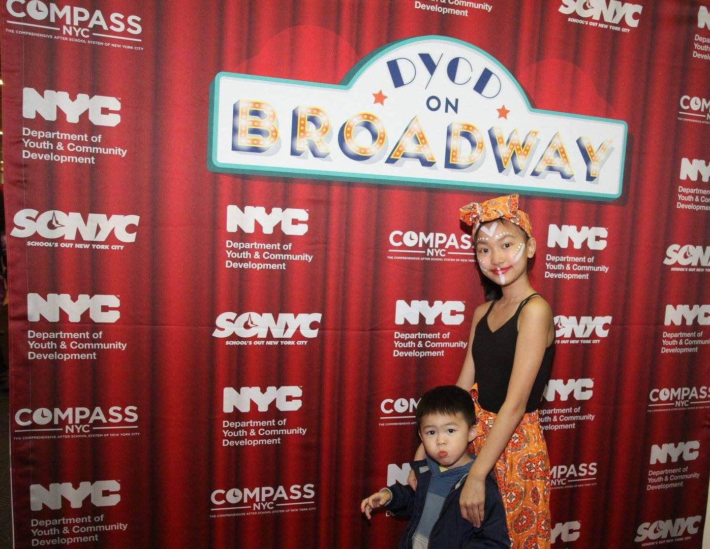 A young girl and small child pose in front of a red step and repeat designed to imitate red theatre stage curtains. The DYCD on Broadway logo is positioned high and centered on the step and repeat with the DYCD, SONYC, and COMPASS logos repeating in a pattern.