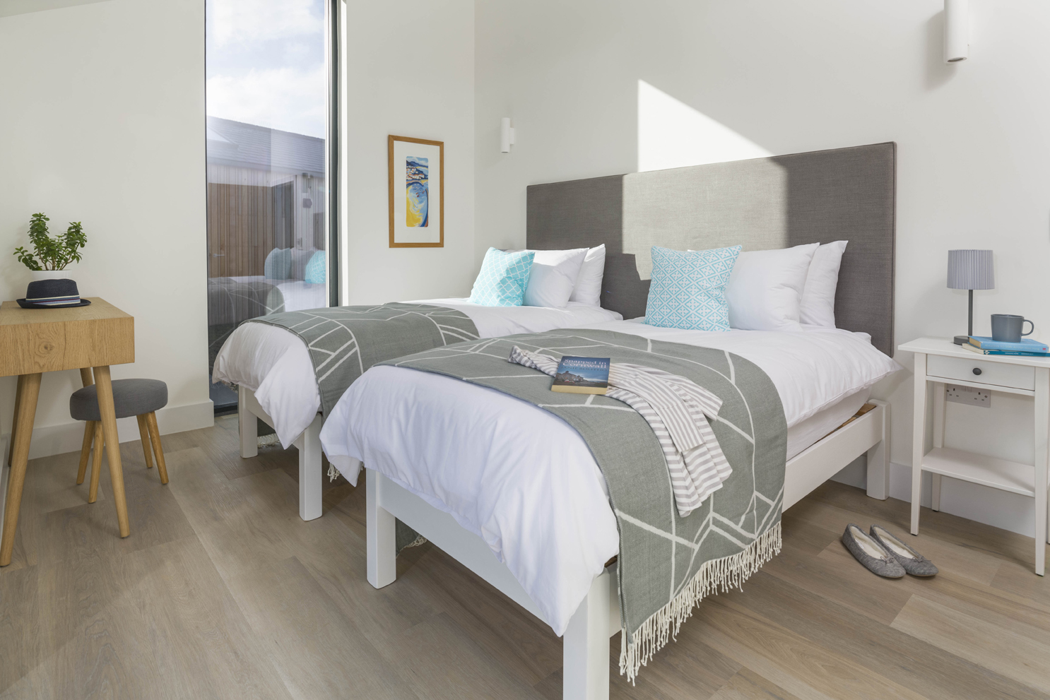 Bright bedroom with single beds and blue, grey and white soft furnishings. Wooden desk with stool and timber flooring. 