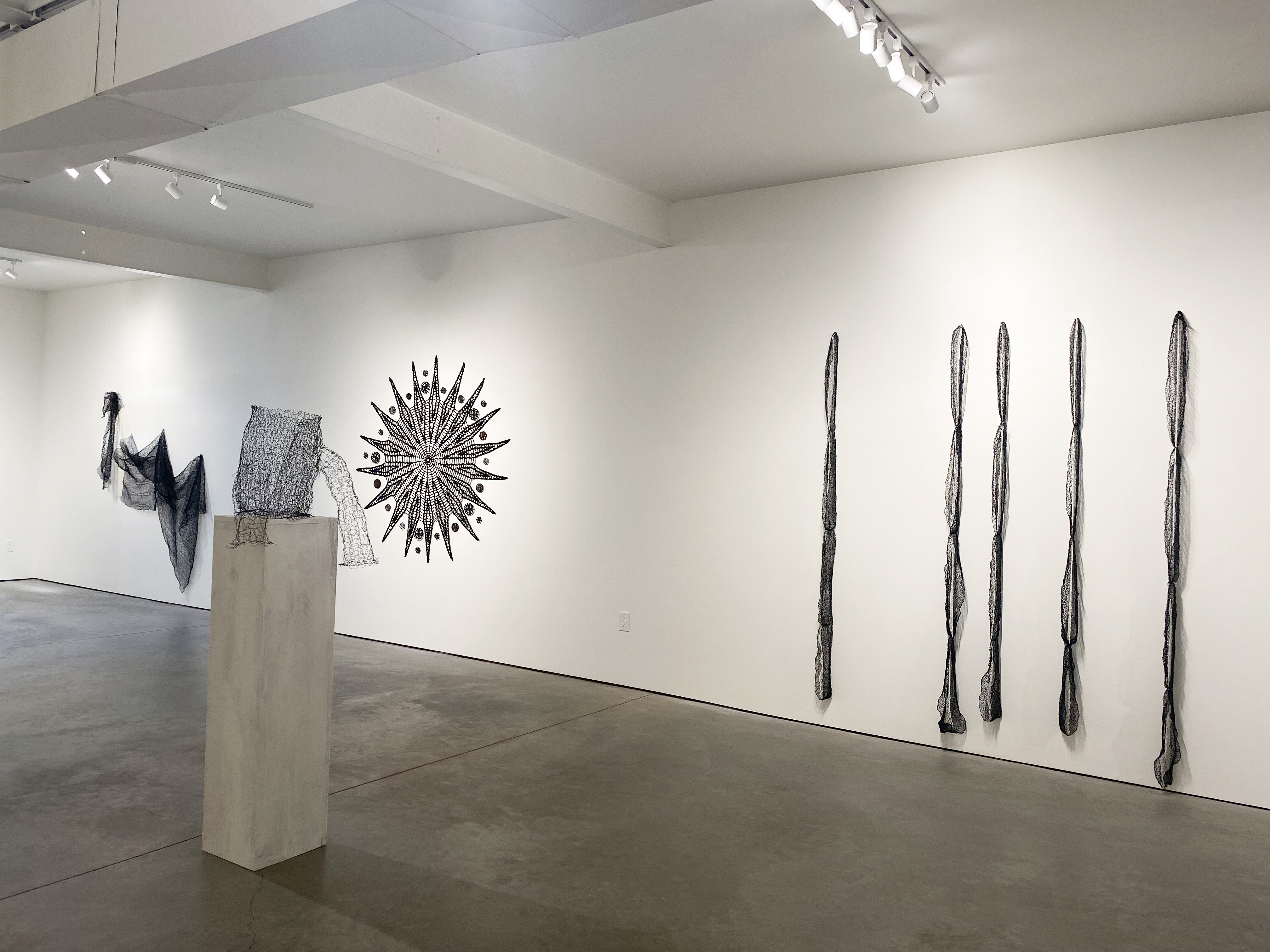 Interior view of a long wall in the gallery Quappi Projects, a brightly lit white wall gallery with various wall-hanging sculptures wrapping around the room. To the right and closest to view is a series of 5 long, thin pieces of hanging black fabric in a row.
