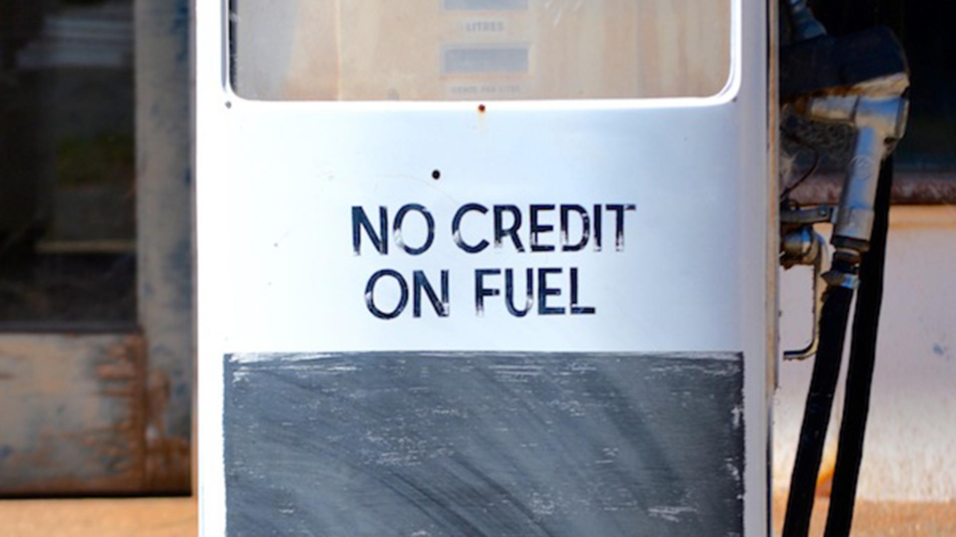 ID: Close-up of video still from 'No Credit'. An old Mobil petrol pump station with worn text reading "NO CREDIT ON FUEL". The lower half of the pump station has been painted with blackboard paint, which is also distressed.