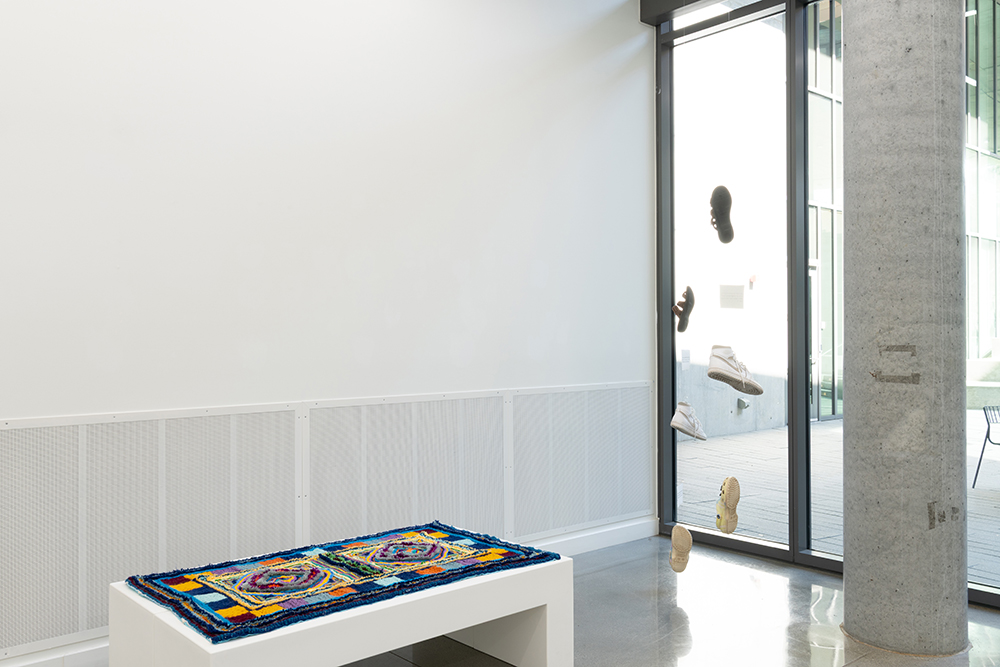 A rug on a bench or platform and shoes and papers hanging on two strings in a gallery space.