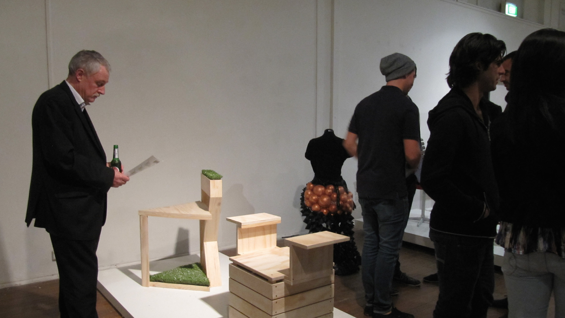 ID: A crowded exhibition opening. A man stands next to a series of modern hand-made chairs.