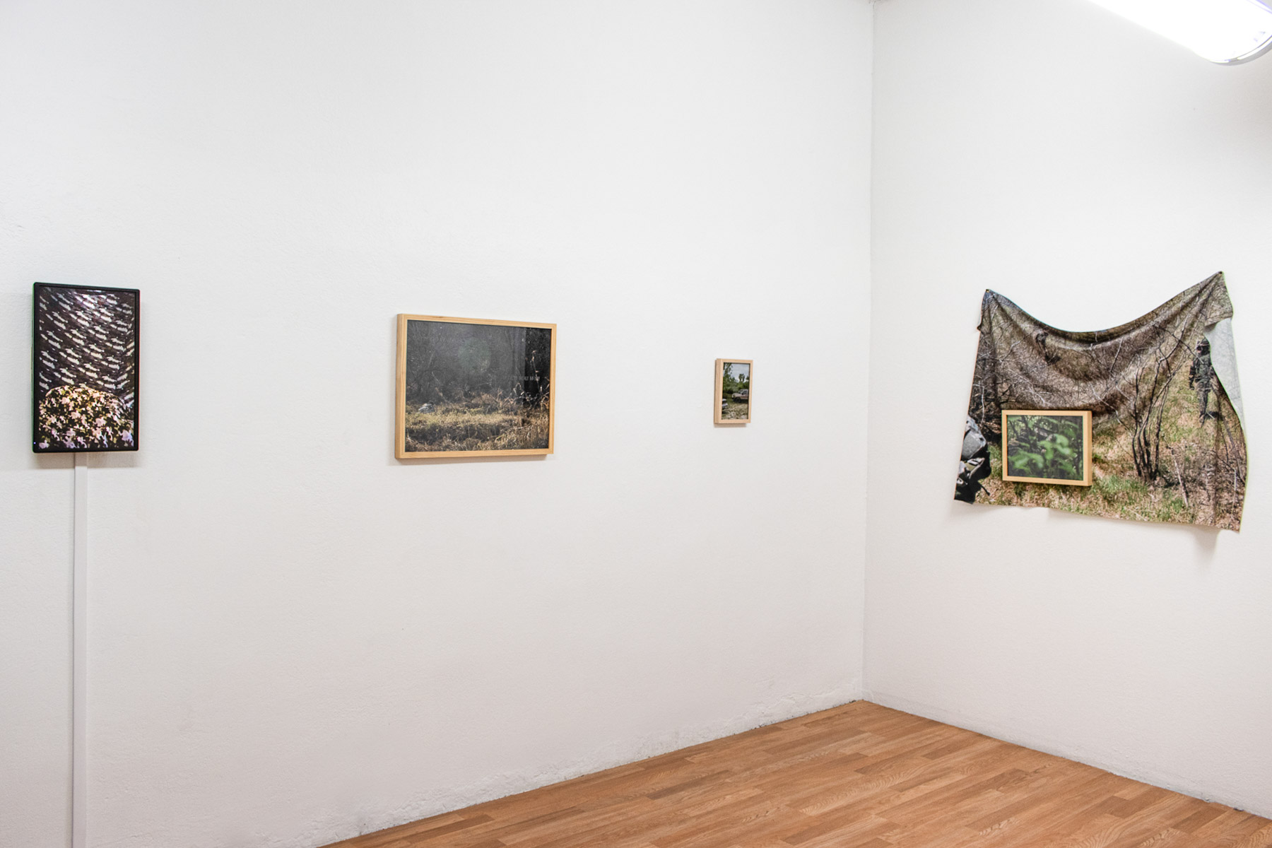 A corner with white walls and a light wood floor. On the walls from left to right are a video monitor, two framed photos with scenes of grass, trees, people in camouflage, and a truck, and a draped fabric photograph of people in camouflage with a smaller framed photo hung overtop of it.