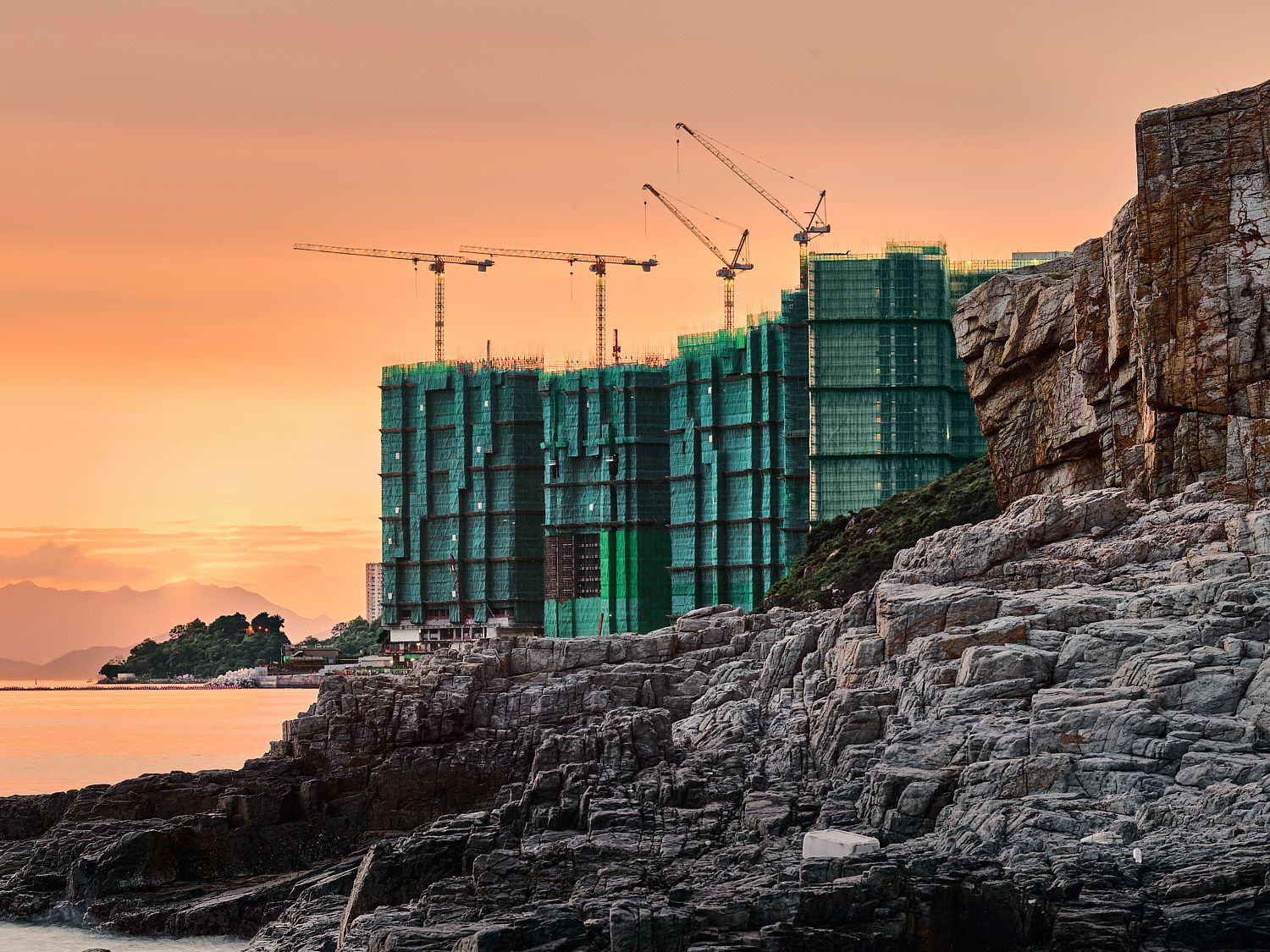 Scaffolded construction site of a large property development in orange light, rocky coastline, cranes, Hong Kong waters