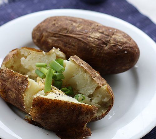 A simple picture of two baked potatoes on a plate with one potato open with chives placed on top