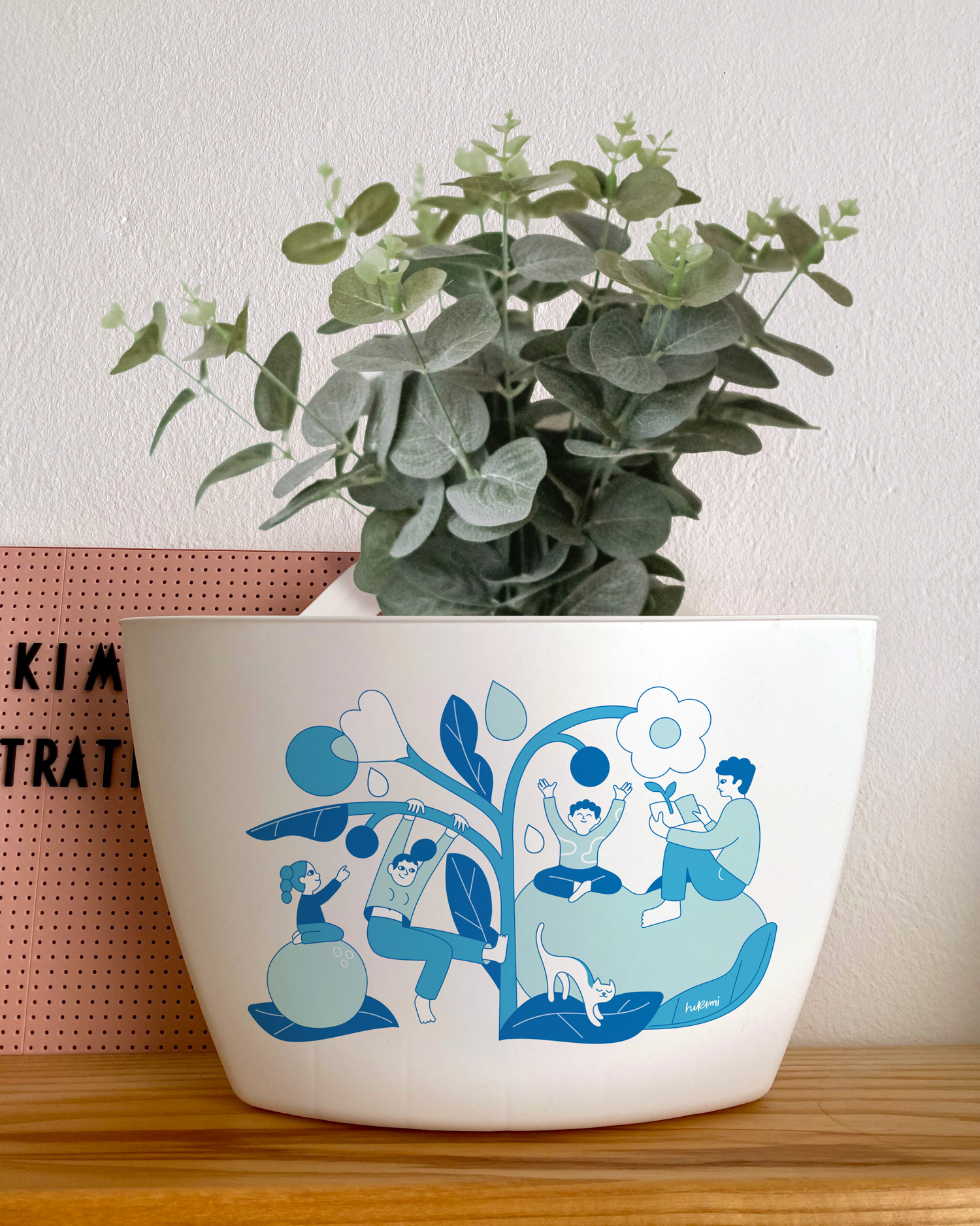 A white planter with an illustration in shades of blue featuring people interacting with a stylized plant. The scene includes a person reading on a leaf, another meditating, and a child climbing a branch. A cat is also present, adding a playful touch. The illustration, designed by Hikimi, conveys a sense of balance and connection with nature.