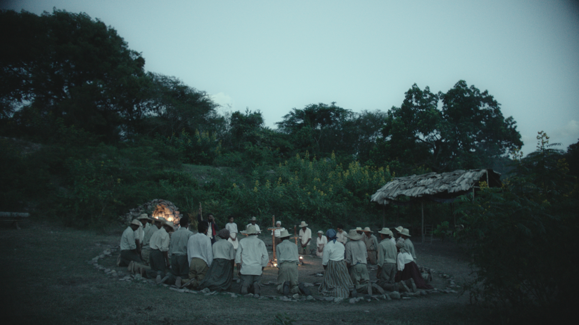 Image Description: A photo of a group of people kneeling in a circle around a circular campfire that has been marked out with a large ring of stones. They are wearing tan and white clothing and most of them wear some type of head covering, whether a har of head scarf. Their heads are slightly bowed. A lush forest can be seen in the background.