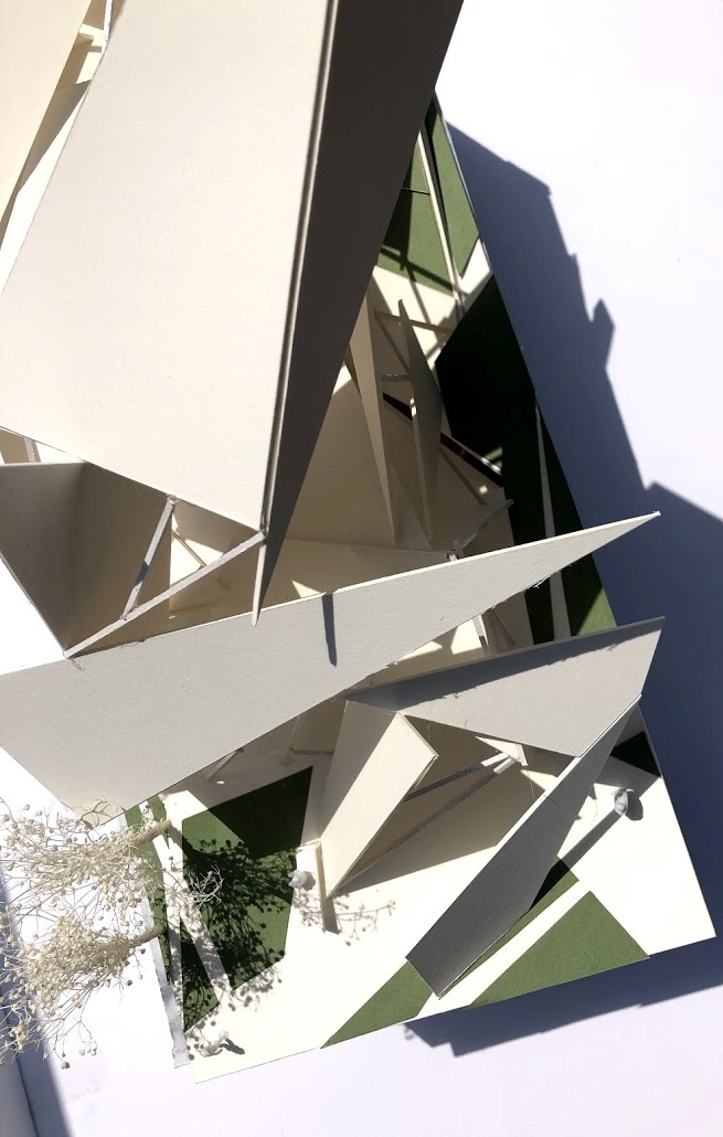 a photo of the model made out of white chipboard - it looks like origami delicately floating in place. green spaces are represented with green textured paper, along with small model trees. scale people are also visible.