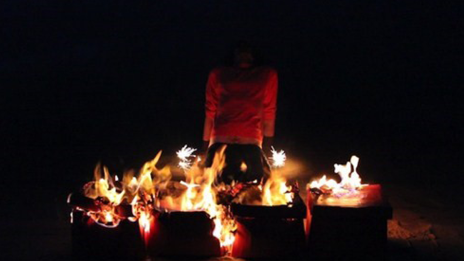 ID: A blurred image of a person kneeling behind a ring of boxes. The features on the person's face are not visible. The person is wearing an orange shirt. In the foreground, the top of the boxes have been lit on fire. Either side of the figure in the background are two lit sparklers. The background is in shadow.