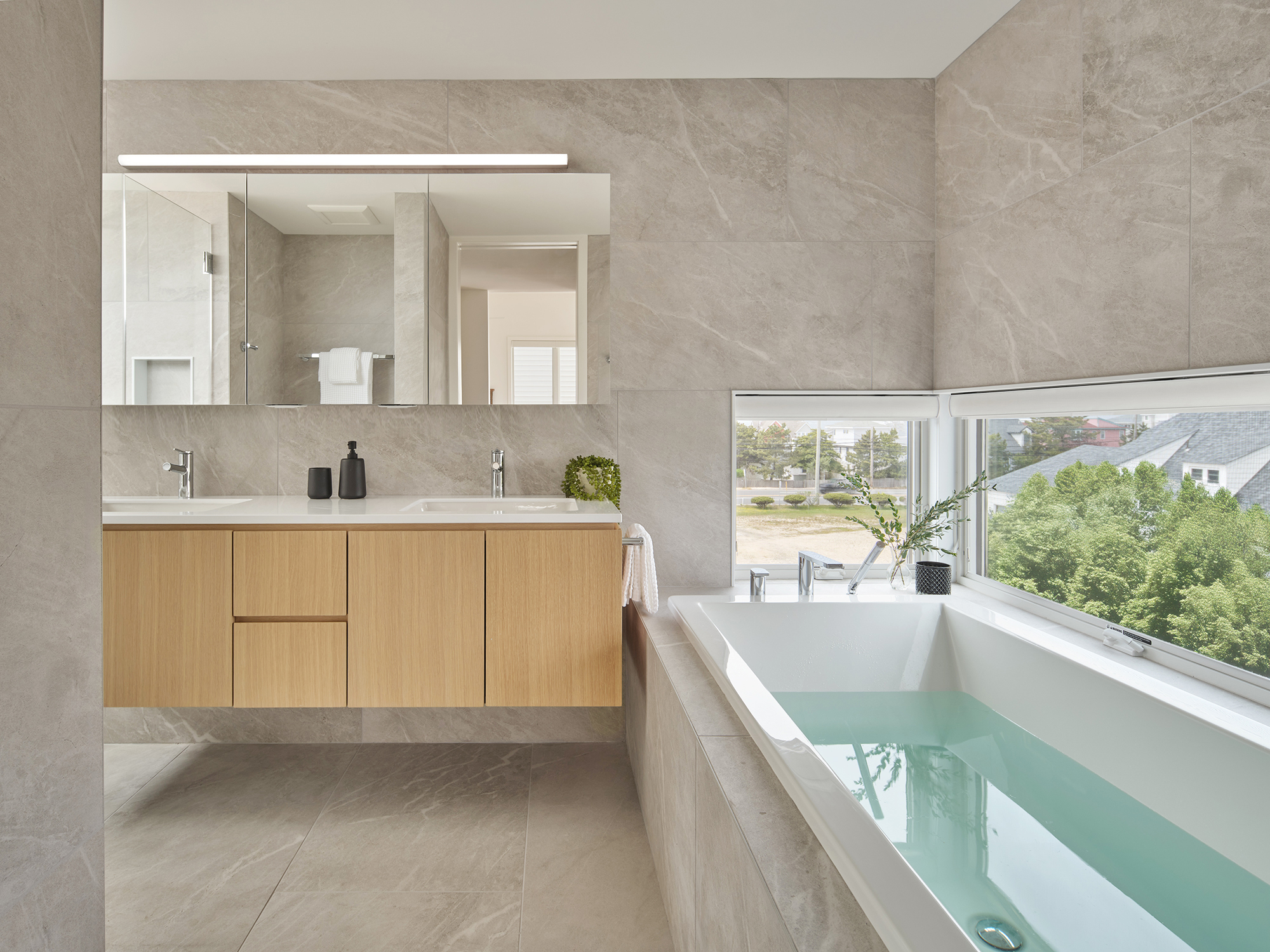 Architectural render of the Gap House Main Bathroom, showing a modern soaking tub beneath a window, a light wood double vanity, and gray large-format wall and floor tiles.
