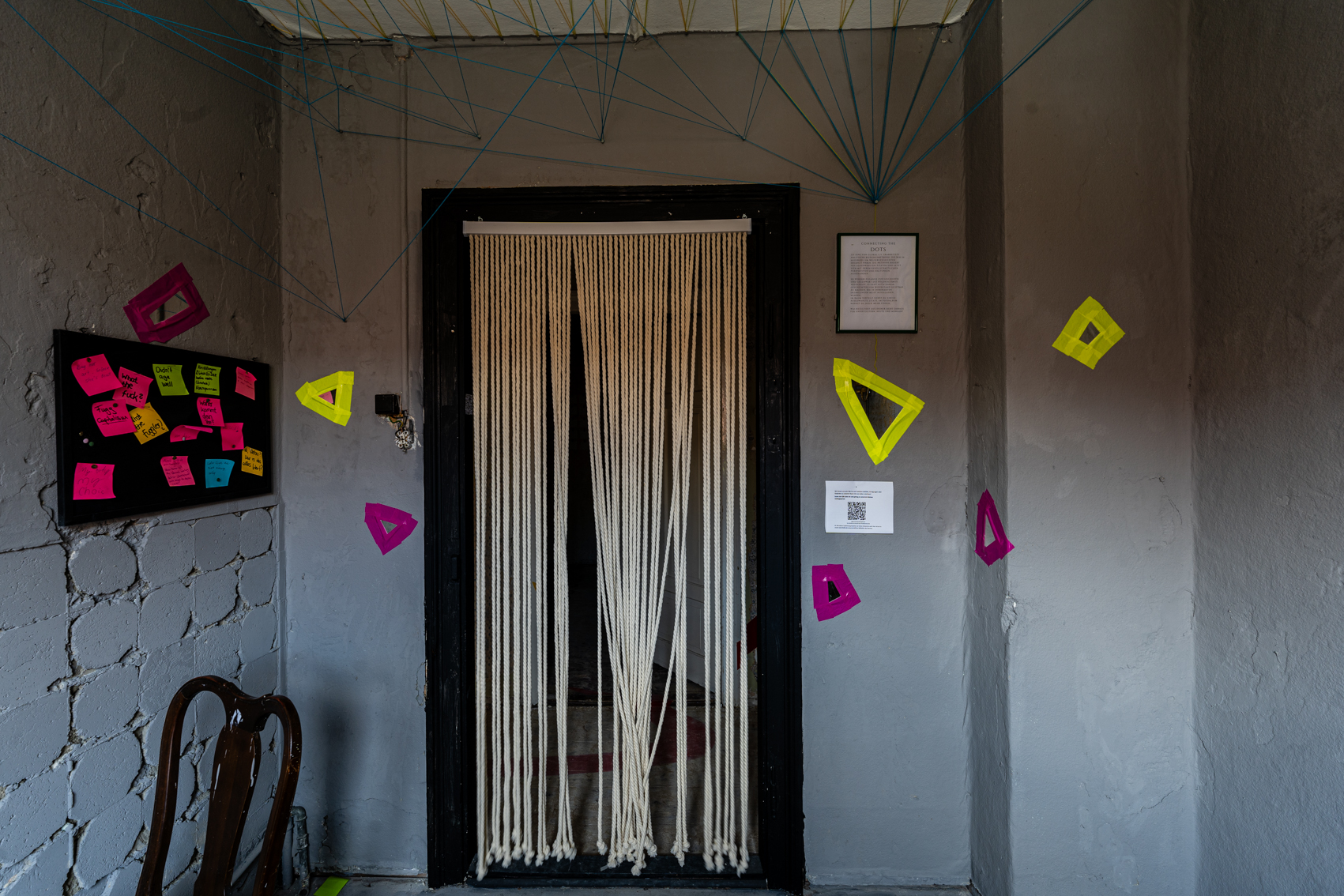 Bild 2 zu Ein Schritt zur&uuml;ck nach vorn: Blick im Raum stehend auf den Ausgang. Schwarzer T&uuml;rrahmen mit Vorhang aus vielen Stoffkordeln. An der Wand vereinzelte Dreiecke aus gelbem und pinkem Klebeband. Links an der Wand ein schwarzes Brett mit Post-its.