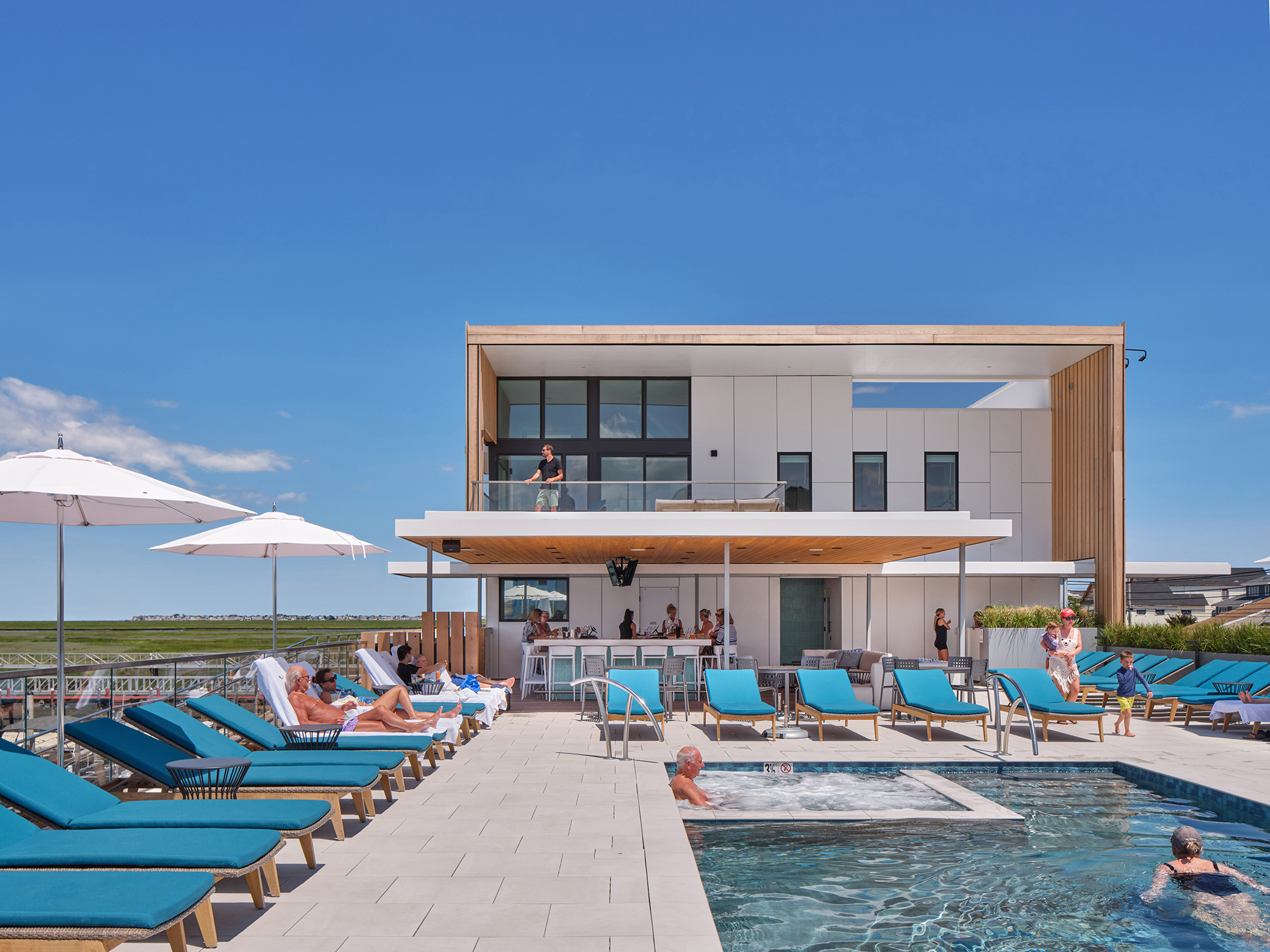 Exterior view of the Brigantine Paddle Club elevated pool deck, showing sunbathers, a small spa, blue lounge chairs, and the modern white and wood-clad club building in the background.
