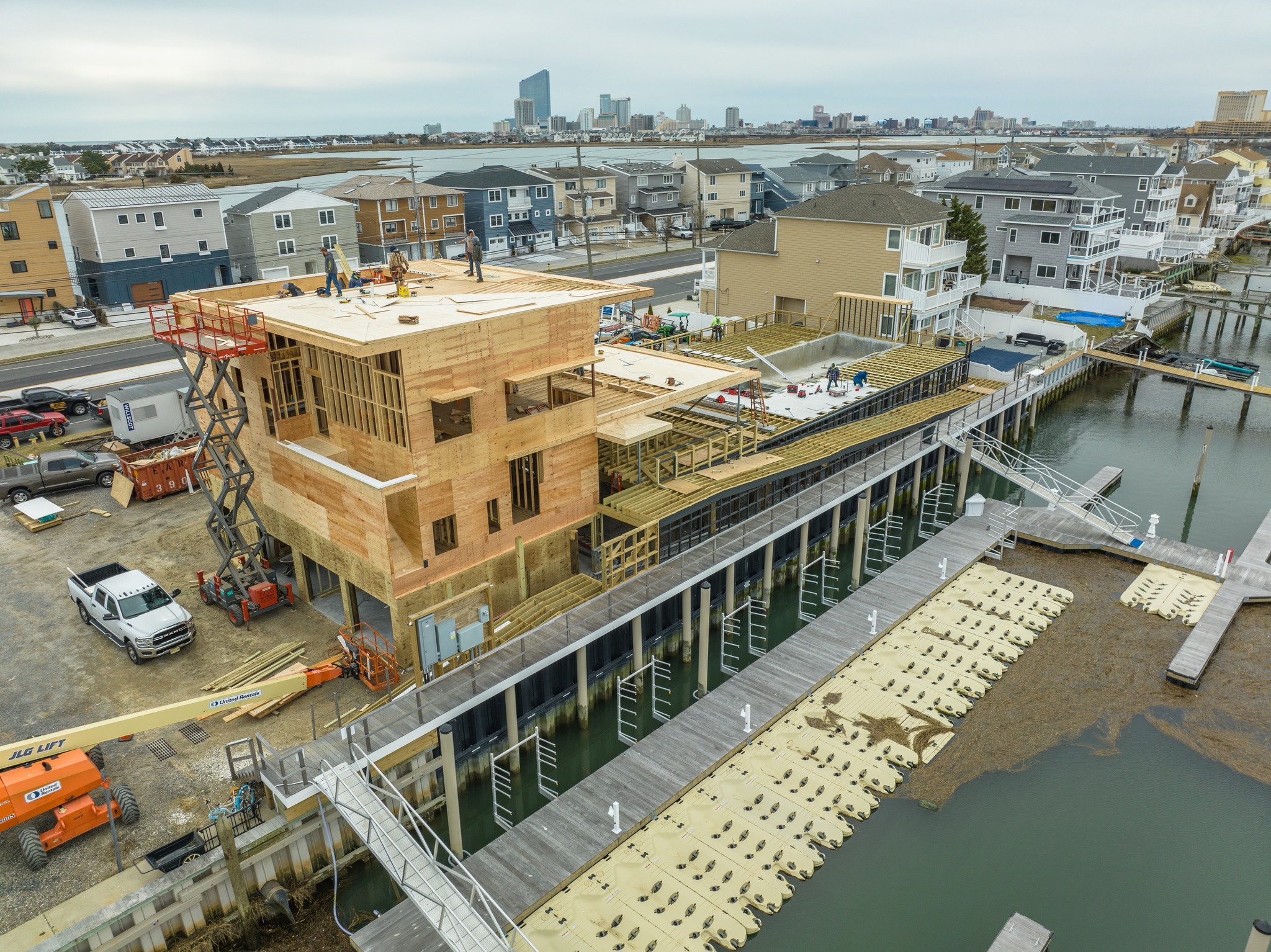 Aerial drone photo of the Brigantine Marina Paddle Club construction, highlighting the building's framing and the elevated deck structure, with a distant city skyline visible.