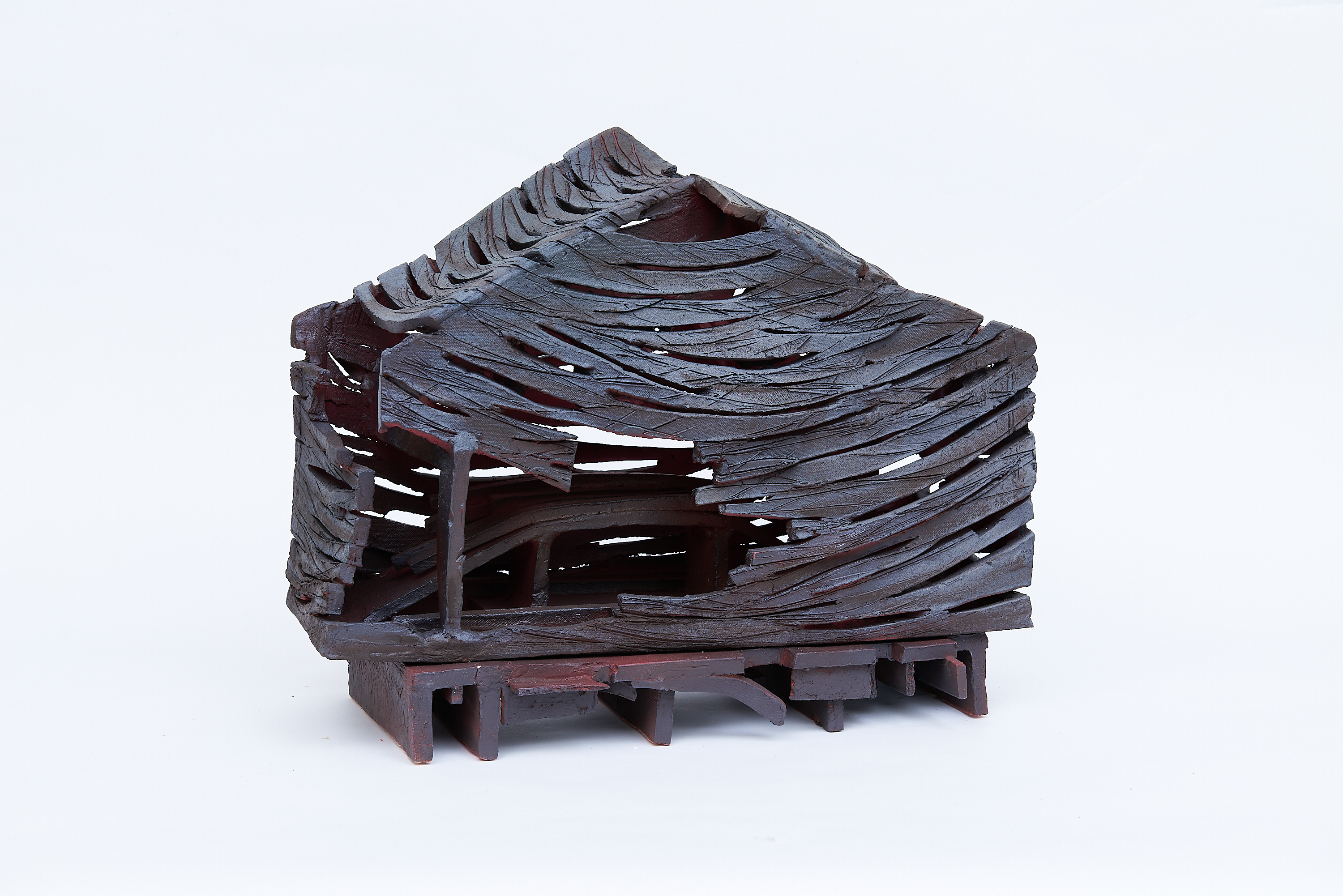 Lydia Thompson, Tumble, 2023. Ceramic, 22 xx 17 x 6.5". A dark brown house made from slashed clay strips with a hole in the roof, middle left, and far left resting on a pallet of the same color