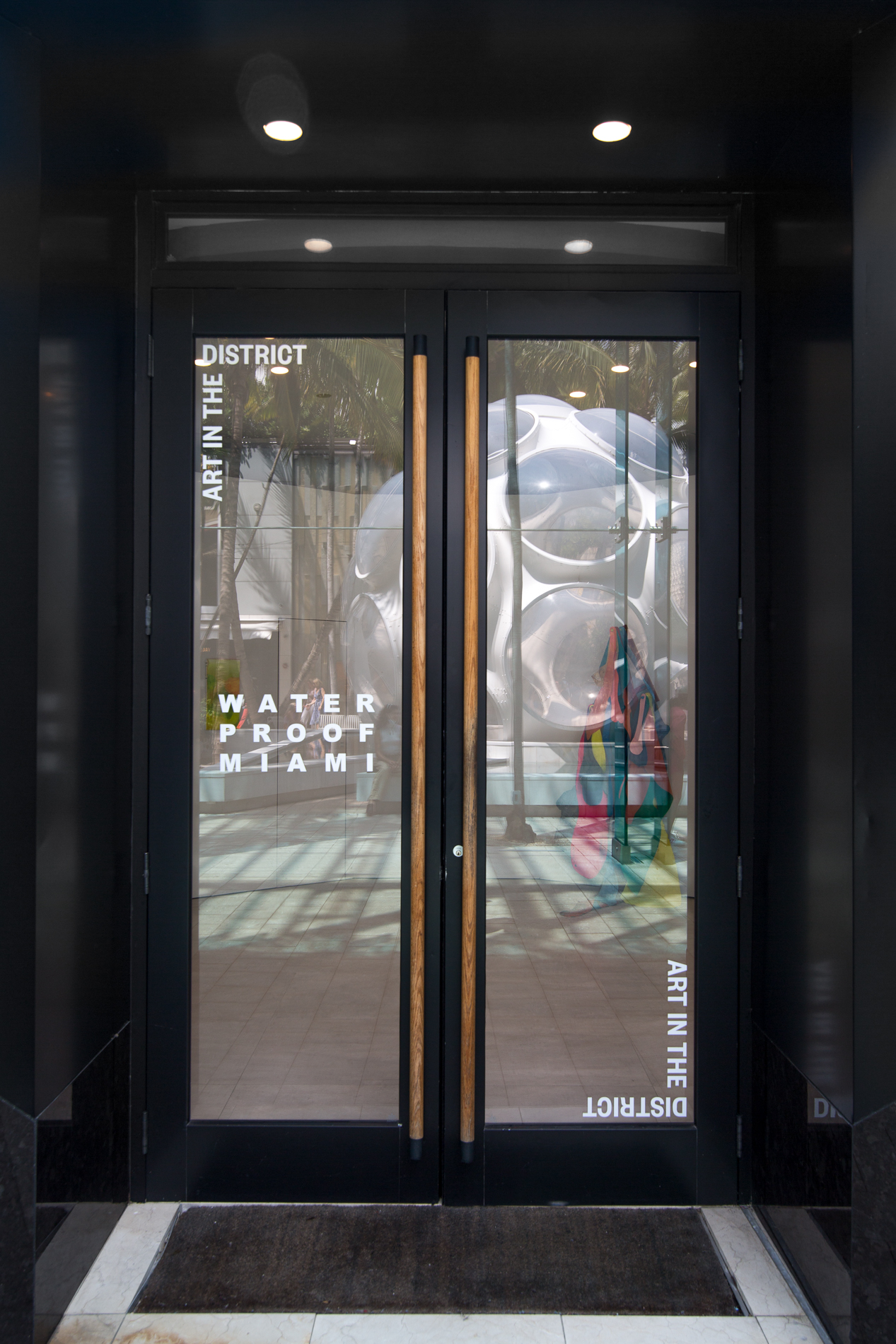Nadir Sourgi, Installation view of Metonym at Bas Fisher Invitational as part of Waterproof Miami, Photo by Monica McGivern (2022). Two french doors, outlined in black with wooden elongated pull handles, have on diagonally opposite corners in white text “Art in the District ''. Centered on the left door reads “Waterproof Miami”.