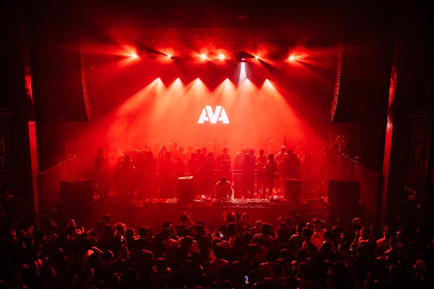 DJ performing at AVA London event under vivid red lights with crowd dancing below AVA logo.