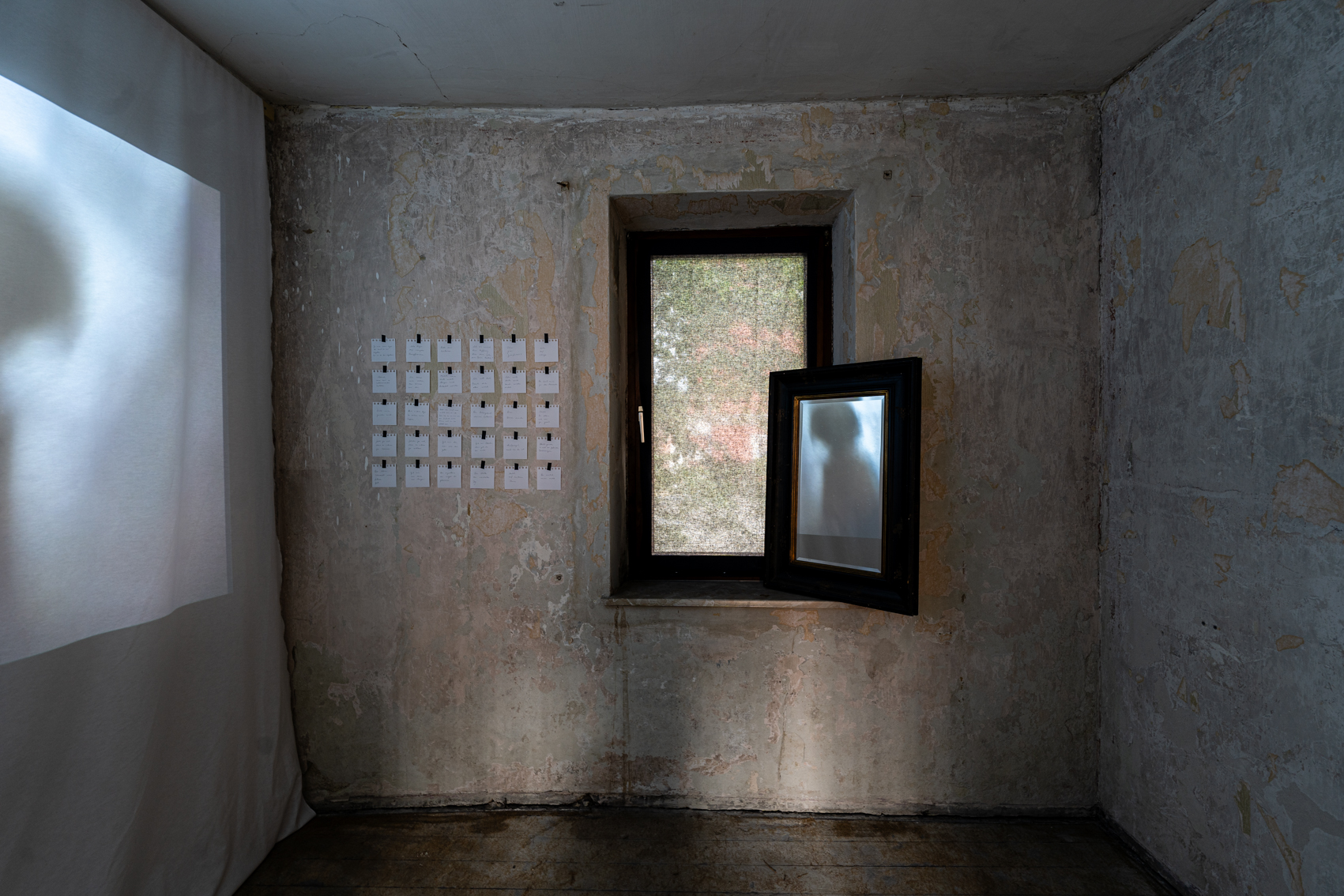 Bild 2 zu An den W&auml;nden der Stadt: Gegen&uuml;ber an der Wand ein braunes Holzfenster. Rechts auf dem Fensterbrett lehnt ein Spiegel, in dem sich das Video des Beamers, das an die linke Wand geworfen wird, spiegelt. Die Tapete des Raumes br&ouml;ckelt ab. Links des Fensters stehen auf einigen wei&szlig;en quadratischen Notizzetteln kurze Gedichte.