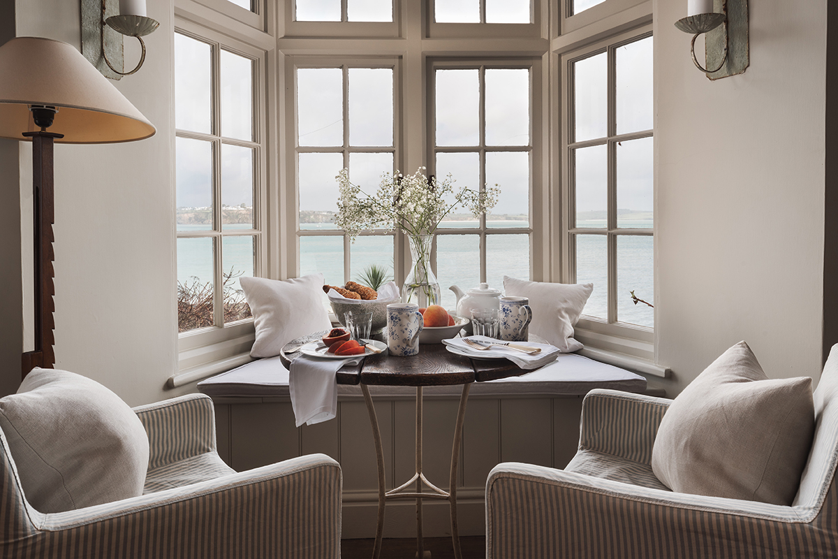 Close-up of two classic armchairs, window seating and table with teatime setting. Overcast sky and sea through window.