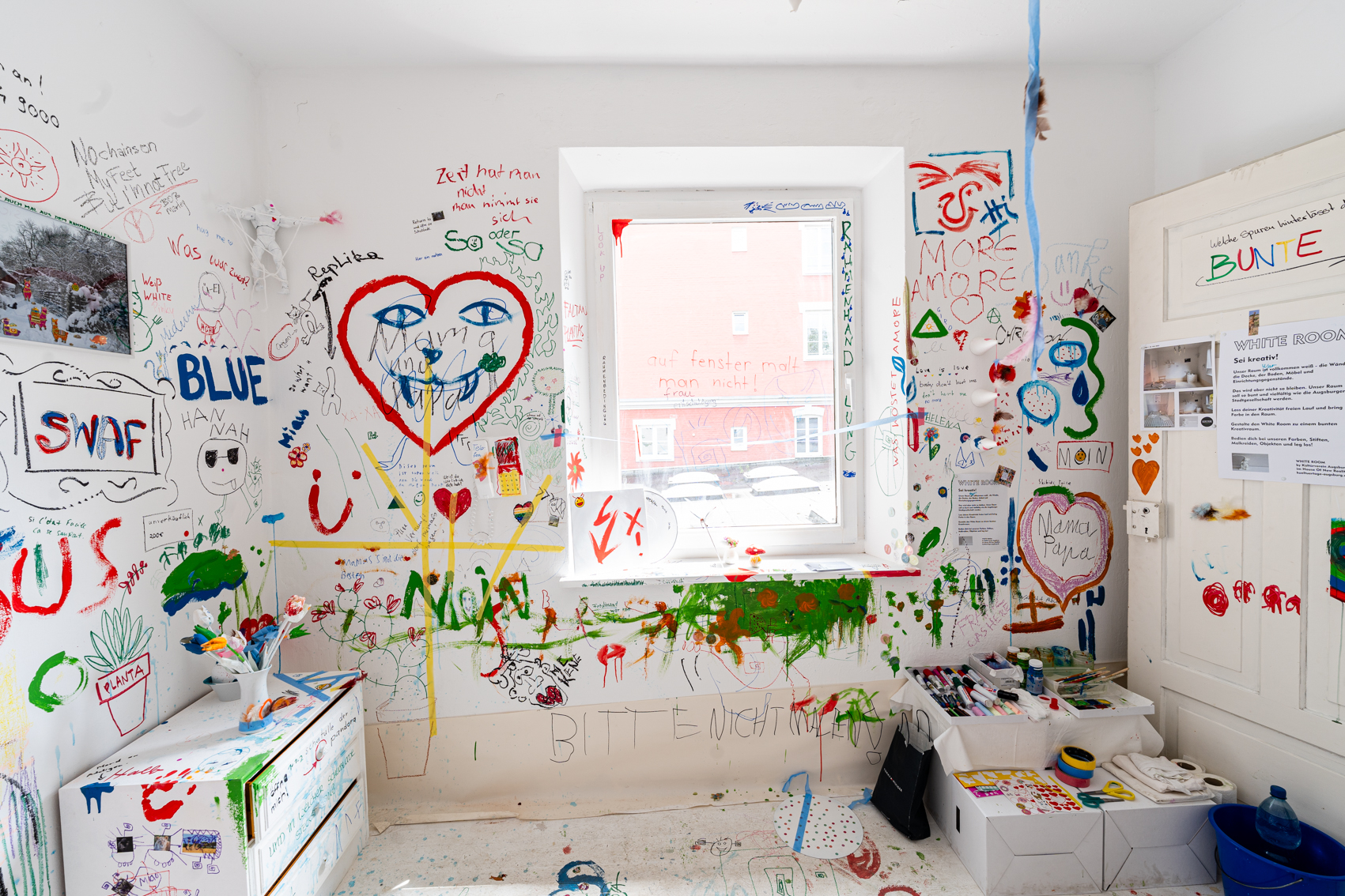 Bild 1 zu White Room: Im Raum stehend gegen&uuml;ber des Fensters. Der Raum ist komplett wei&szlig;. Auf den W&auml;nden sind bunte Malereien, Beschriftungen und Zeichnungen von Besucher*innen.