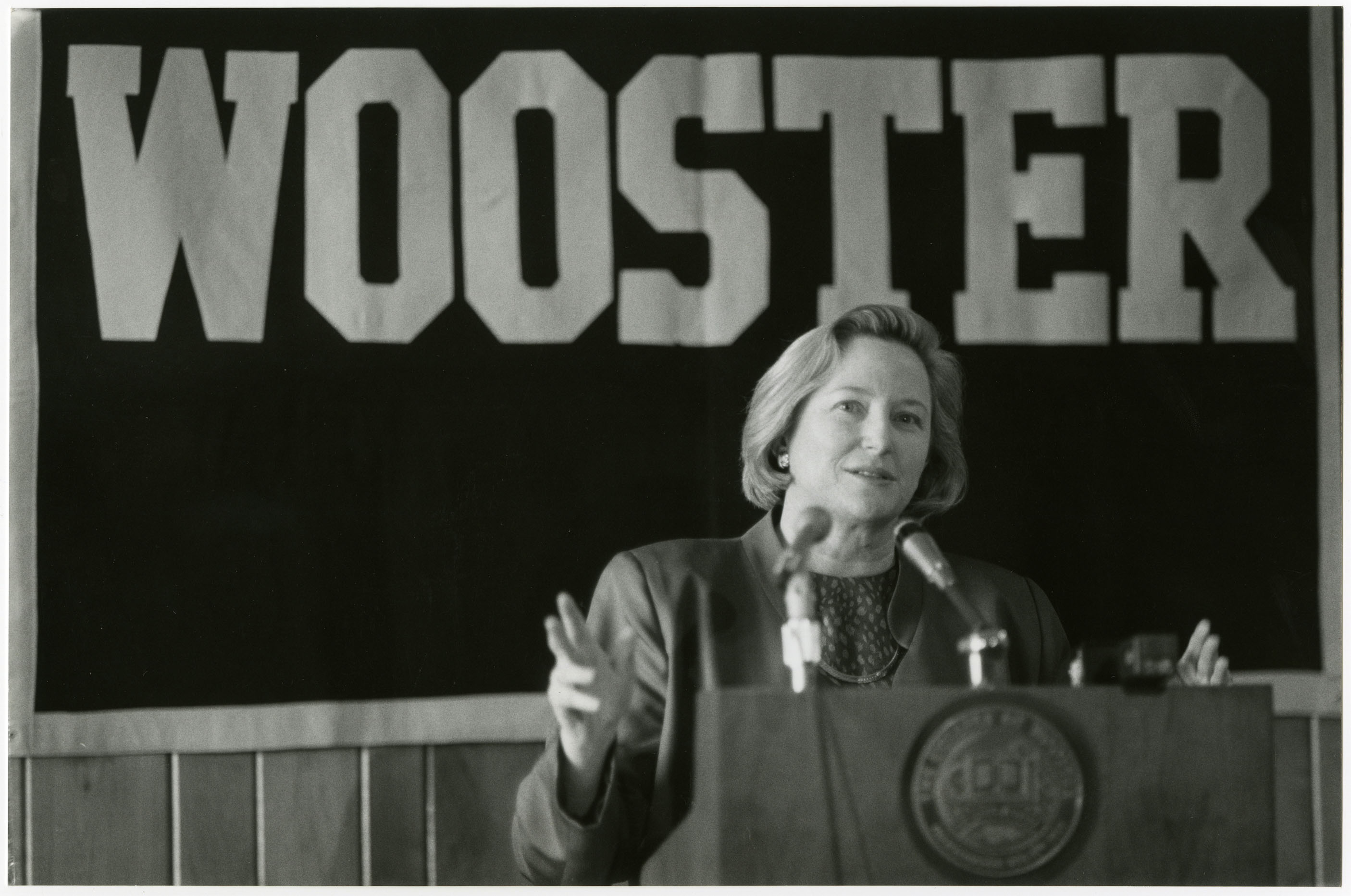 A black and white press conference photograph of Susanne Woods standing behind a podium, with a banner that reads “WOOSTER” hung behind her, and the college’s seal on the front of the pedestal.