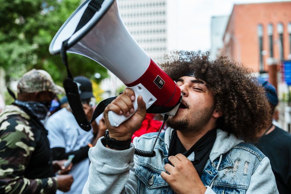 Color photograph of protestor speaking into a megaphone raised to the sky. They are wearing a jean jacket vest and a sweatshirt underneath. Blurry others can be seen behind in various types of clothing: t-shirts, camo and bullet proof vests, ball hats, and helmets.