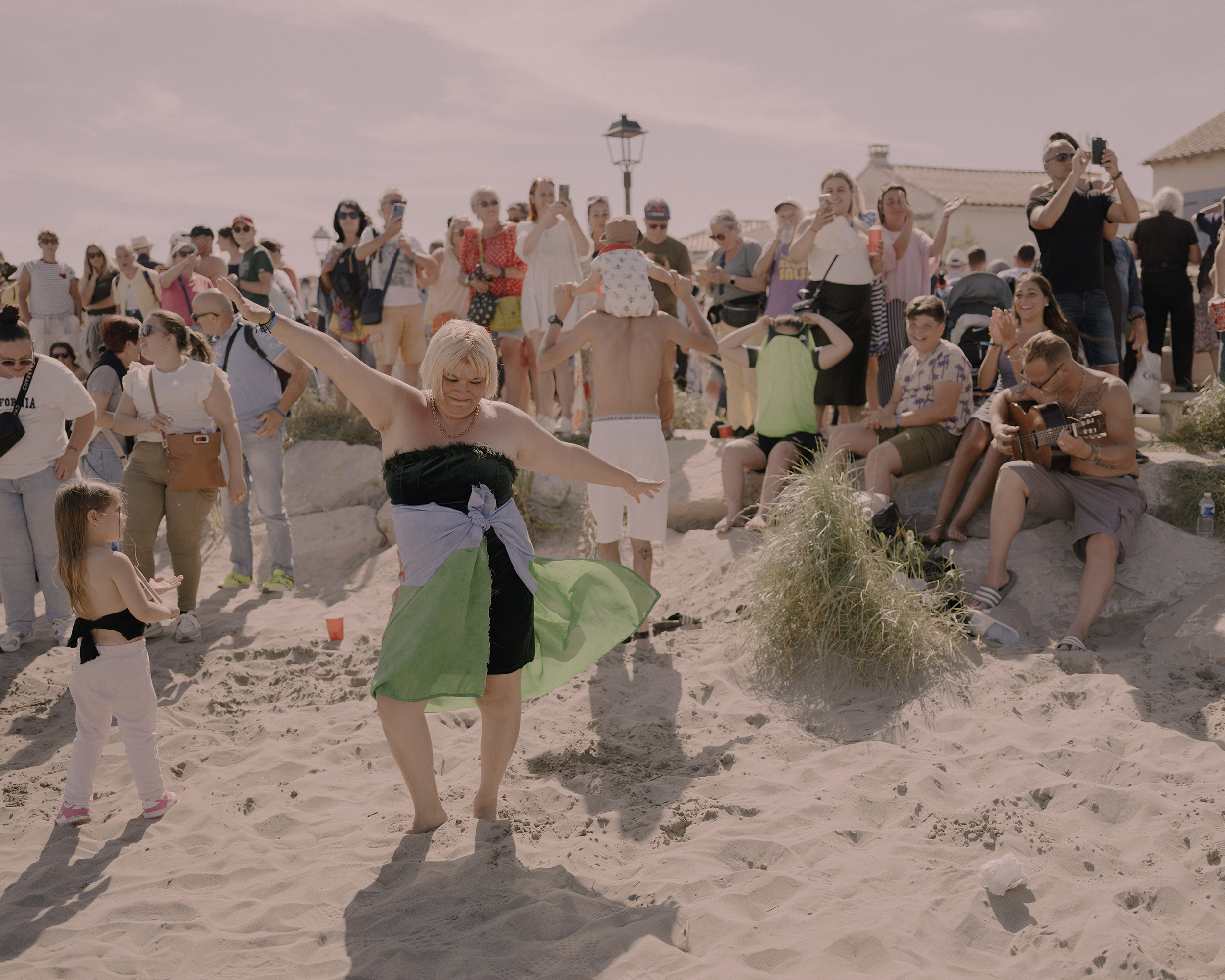 A woman wrapped in a Romani flag dances on the beach as her family watches