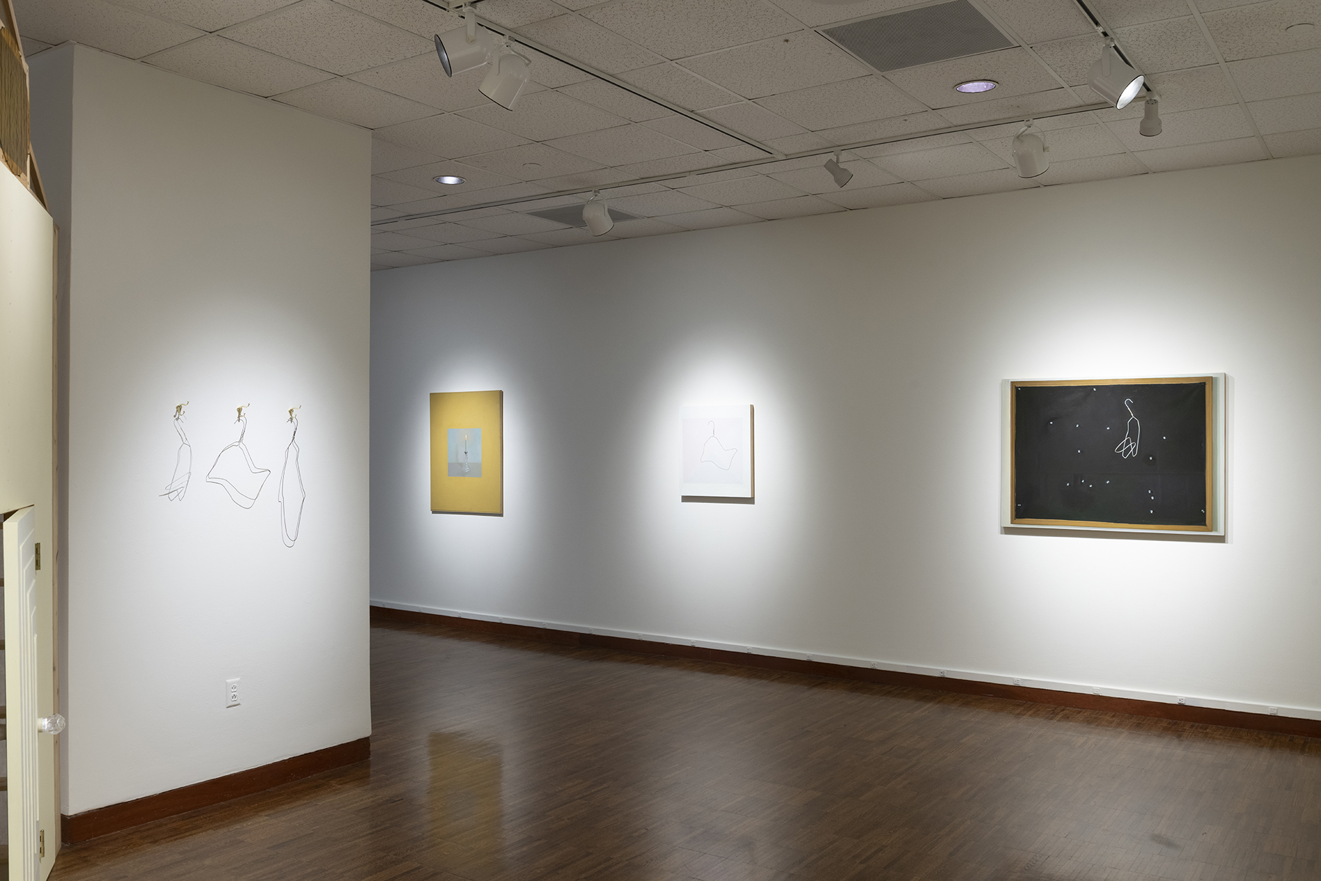 Art exhibition in a white wall gallery space. On a wall to the left appears to be three wire hangers bent into various organic shapes, hanging from brass hooks on the wall. To the right, three still life paintings hang on the wall.