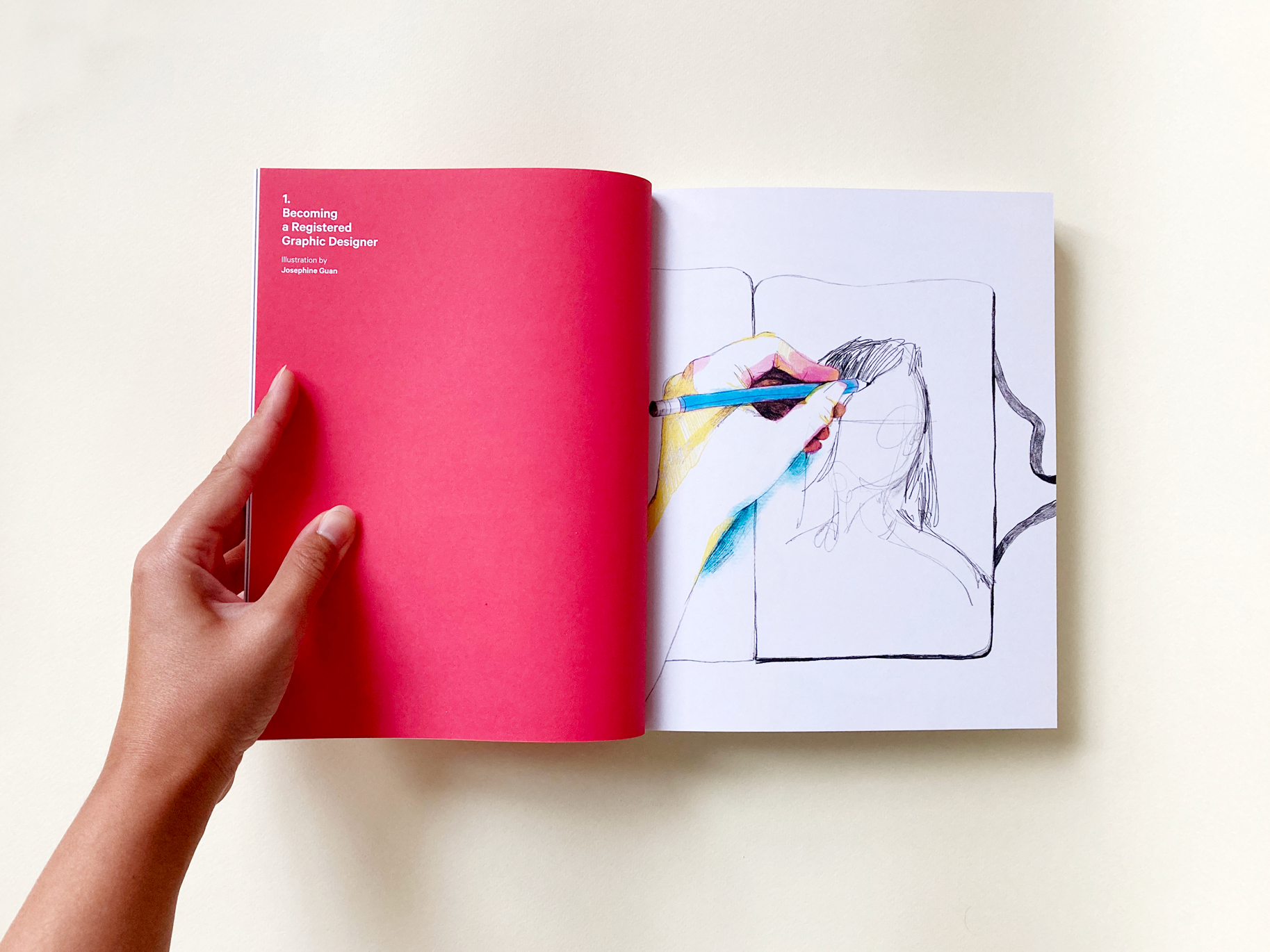Photo of a tanned hand holding open a thick perfect-bound book. The left page is fully red, with white text that reads' 1. Becoming a Graphic Designer, Illustrated by josephine Guan'. On the facing page is a pencil illustration of a hand sketching a portrait in a sketchbook. The portait is not fully formed yet.