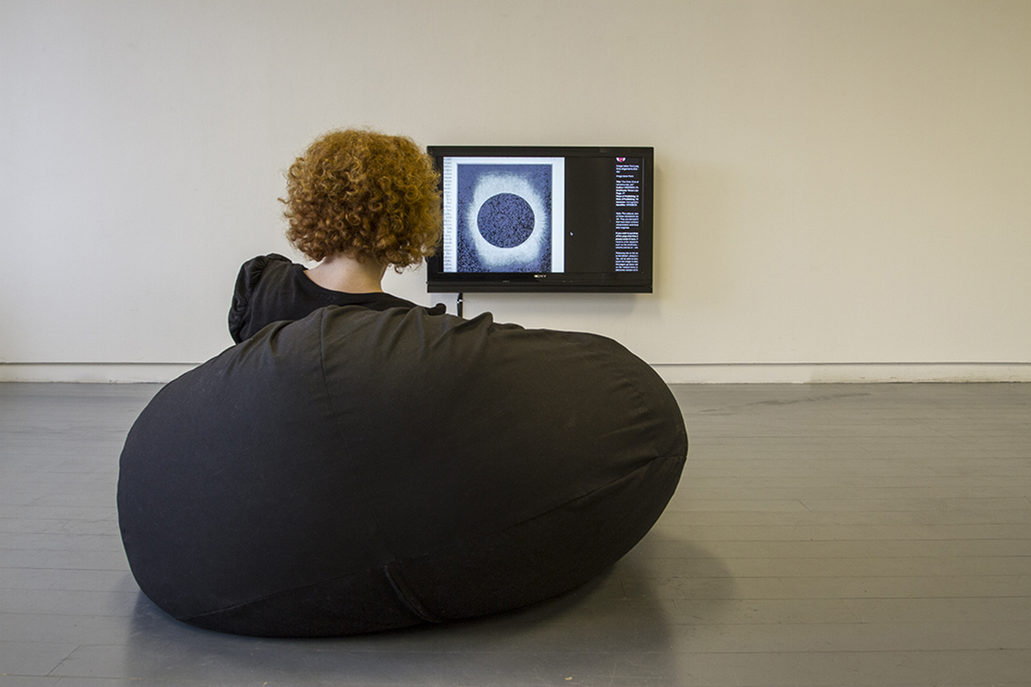 The image shows a detail shot of works in the show After Hours. A red haired woman sits on a black beanbag, looking towards a monitor mounted just above floor level. It is showing the work 'Study for Player Piano (Conlan Nancarrow)', and the screen is filled with a an image of a solar eclipse..