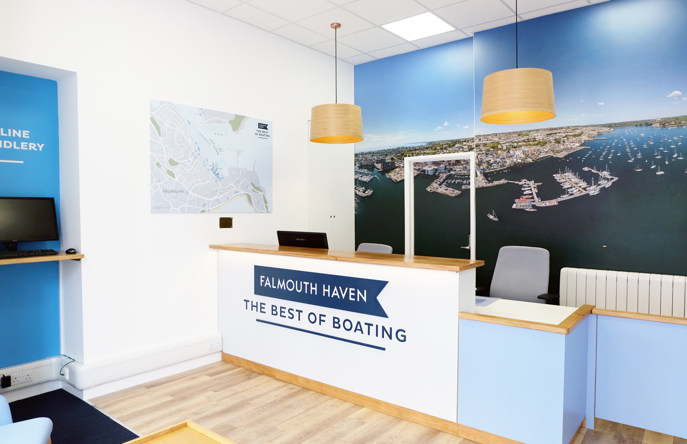 Reception with white and sky blue desk. Logo on front surface and two lighting pendants above. Image of harbour on back wall.  