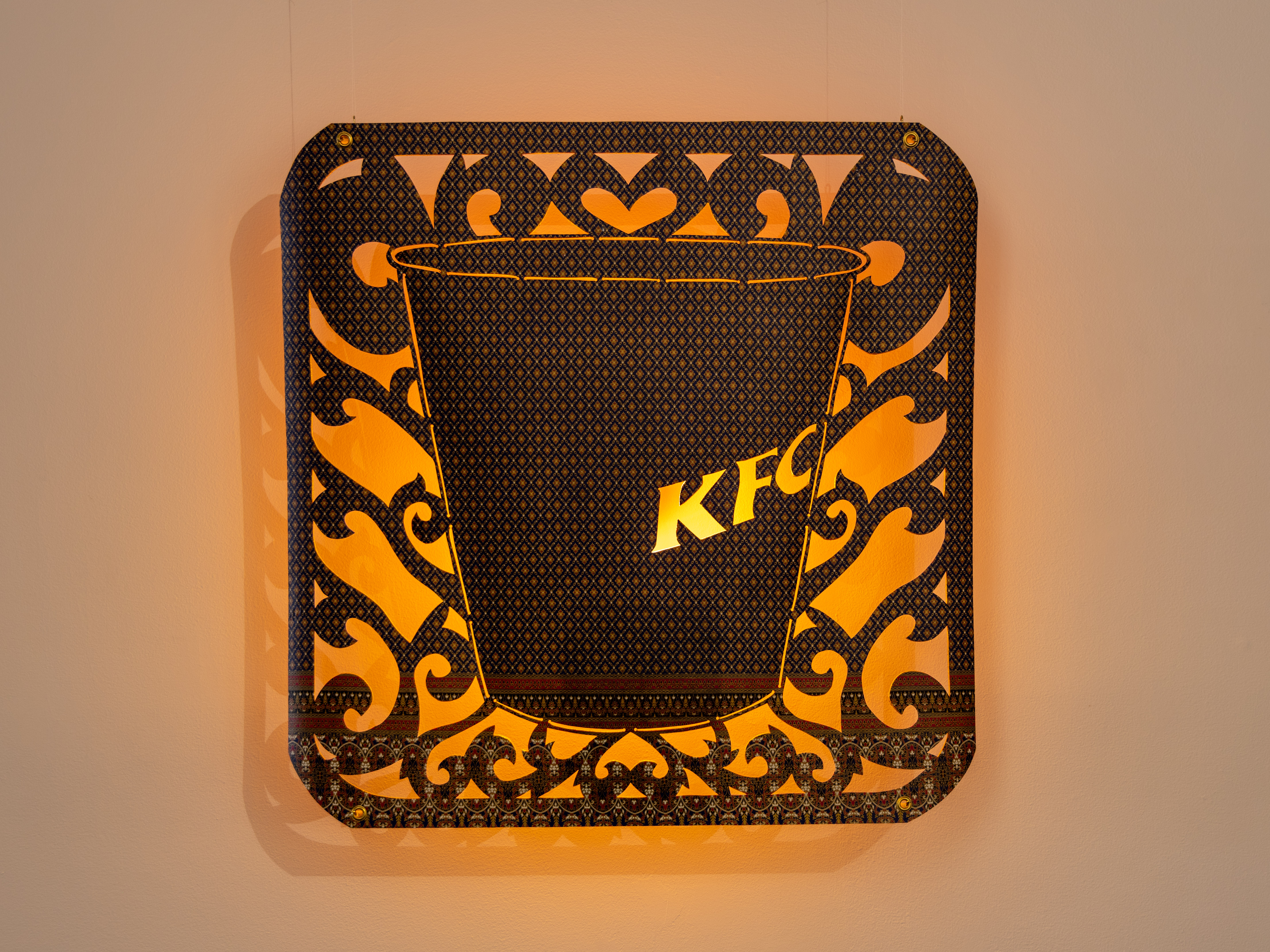 A rounded square piece of fabric is hanging on a wall and is illuminated from behind with a warm glowing light. It is cut in different ways to resemble a Kentucky Fried Chicken to-go container on top of a filigree background.
