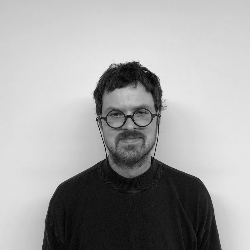 In black and white, a person with round thick classes with neck strings for their glasses. They have brown facial hair, both beard and mustache, and short brown hair. They are wearing a back long sleeve shirt, gazing directly forward with a slight smile.