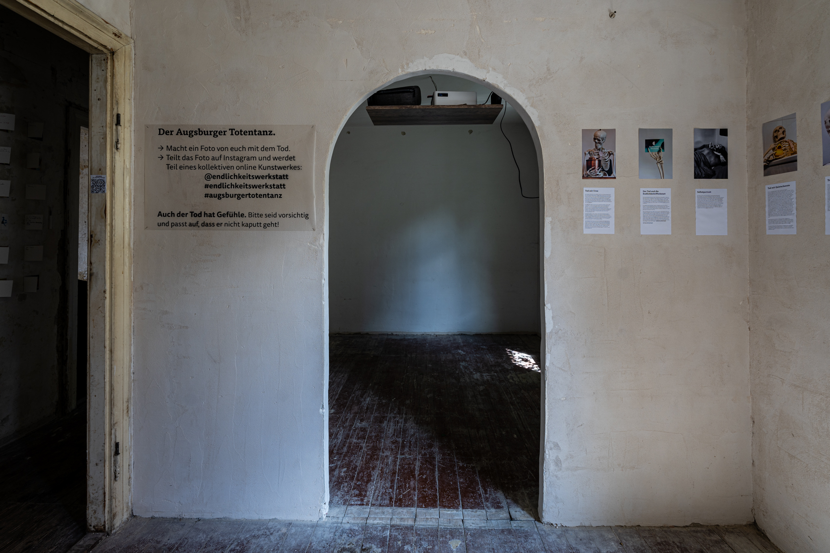 Bild 3 zu Augsburger Totentanz: Blick auf die Wand, mittig ein Rundbogen-Durchgang in den n&auml;chsten Raum.  Rechts an der Wand weitere Bilder und Texte von Skeletten. Links des Durchgangs ein Infotext. Besuchende k&ouml;nnen mit dem Skelett Bilder machen und hochladen.