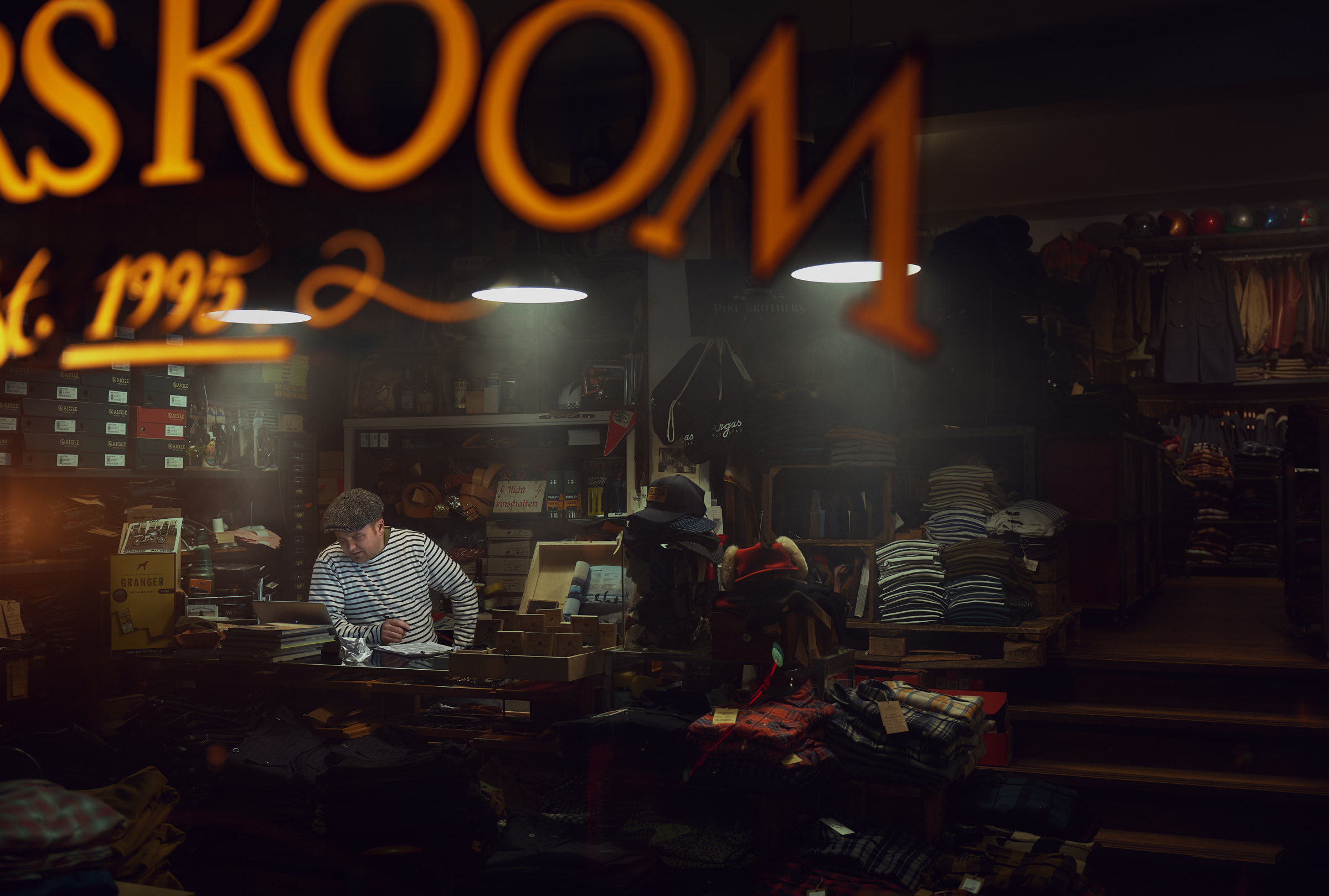 Environmental Portrait eines Mannes sitzend in einem Bekleidungsgeschäft umgeben von klassischer Herren-Freizeitkleidung. Holzfäller- und Karohemden, Lederjacken, ... Stimmungsvolle Beleuchtung.