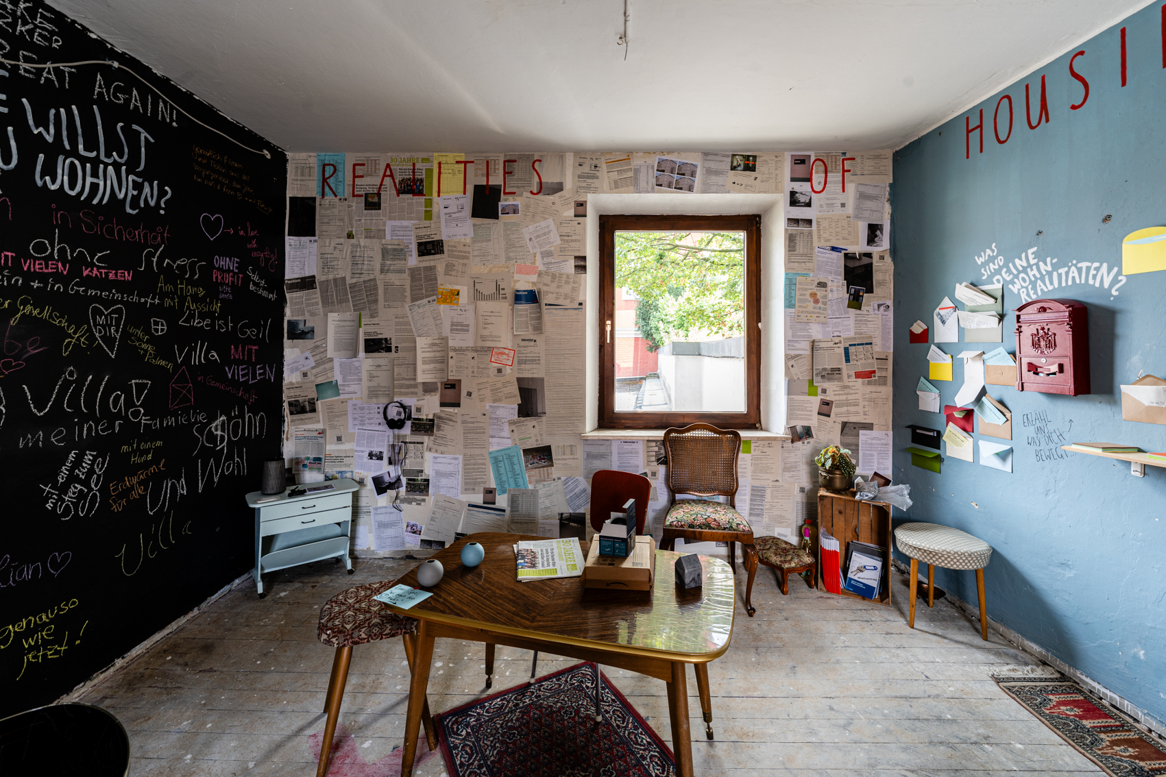 Bild 2 zu Realities of Housing: Blick in Raum mit einem Fenster an der gegen&uuml;berliegenden Wand. Rechts die blaue Wand mit Briefkasten. Mittig ein  niedrieger Holztisch darunter ein roter Teppich. Es stehen vereinzelt St&uuml;hle im Raum. Die komplette Wand des Fensters ist mit Ausdrucken zum Thema Wohnen beh&auml;ngt. Die linke Wand ist schwarz und mit Kreide stehen Antworten von Besucher*innen zur Frage: "Wie willst du wohnen?!"