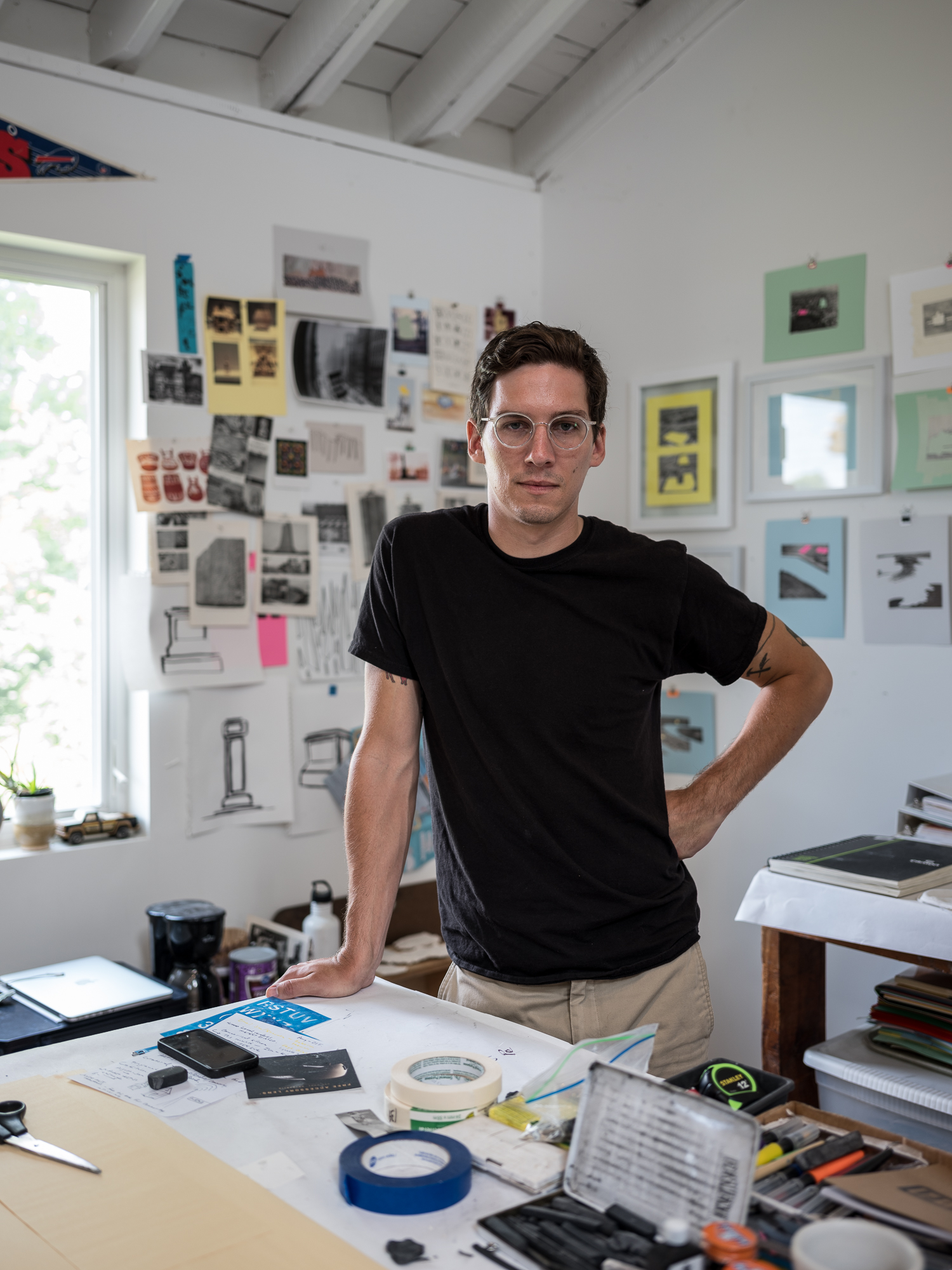 Sean M. Starowitz standing in his studio, just behind a busy desk and in front of a busy wall.