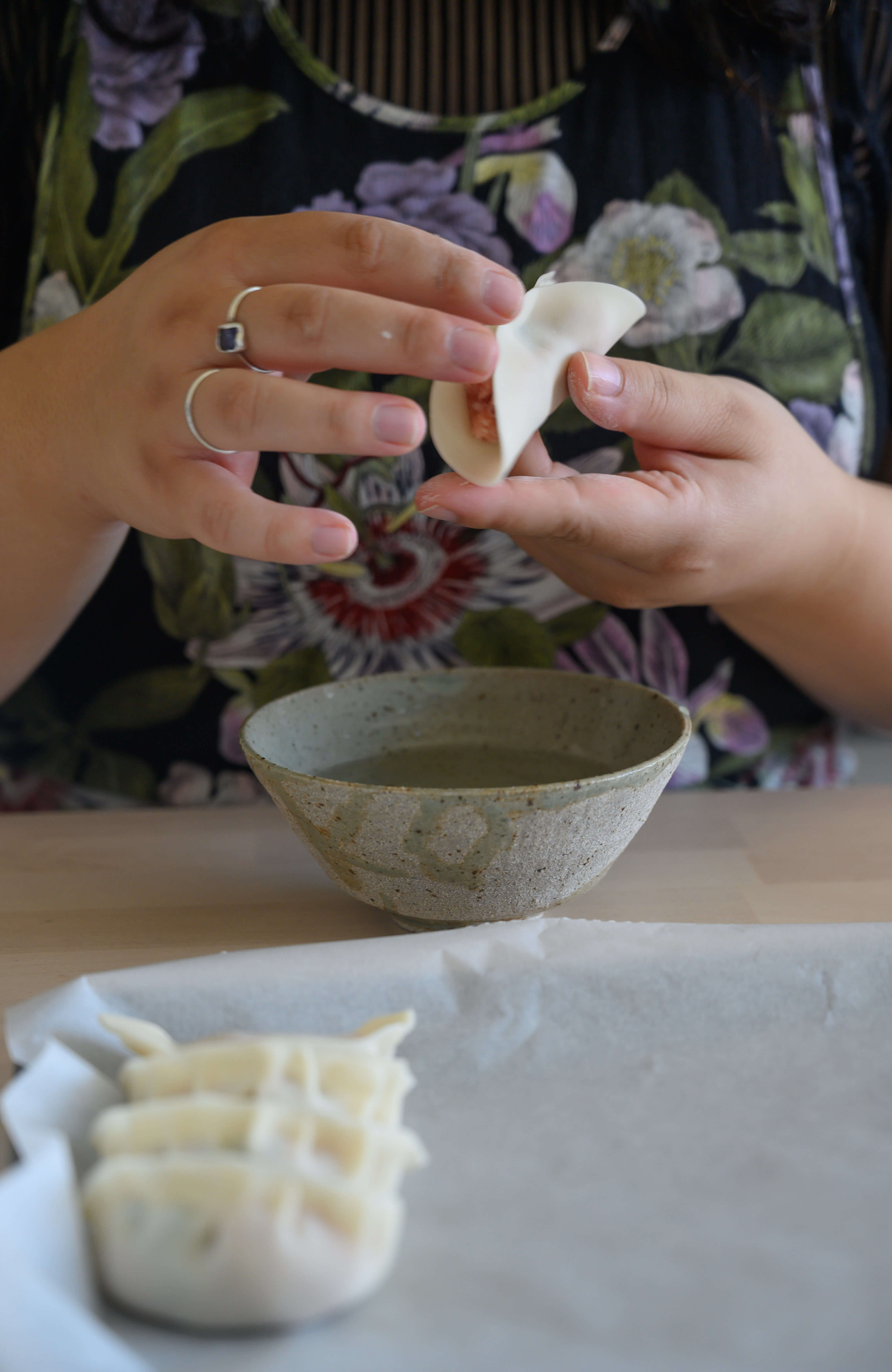 Non’s hands expertly fold a Chinese dumpling. A bowl of water sits below their hands and in front of that is a tray of four folded dumplings.