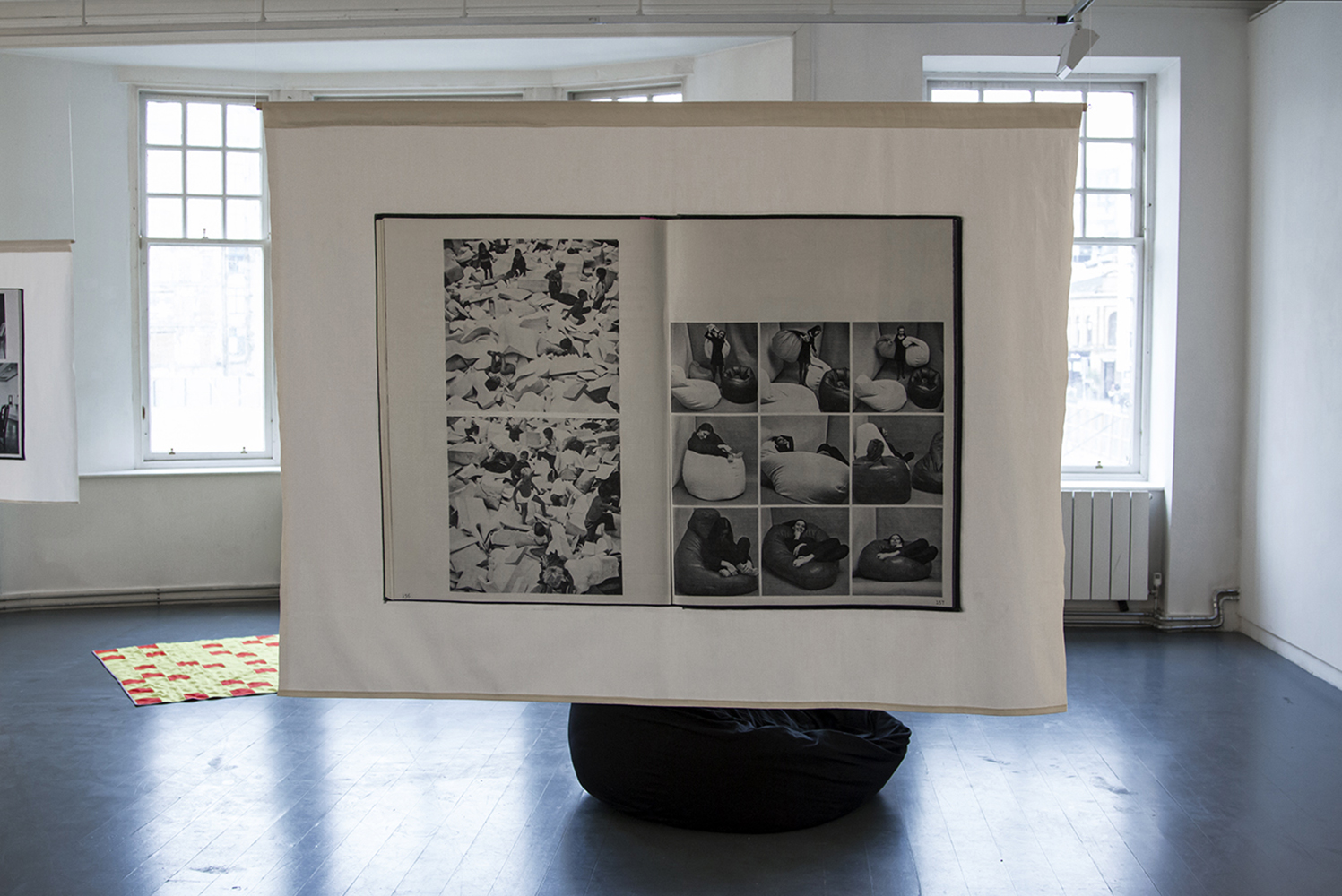 The image shows a printed fabric banner from the show After Hours. The white cotton fabric is printed with a scanned double page spread from the catalogue for 'Information', a landmark 1970 exhibition at MOMA, NY. Both pages feature photographs in black and white. On the right hand page, children play in a soft landscape of foam offcuts and pillows. On the right page, a grid of nine images of a young woman relaxing and playing with a designer beanbag.