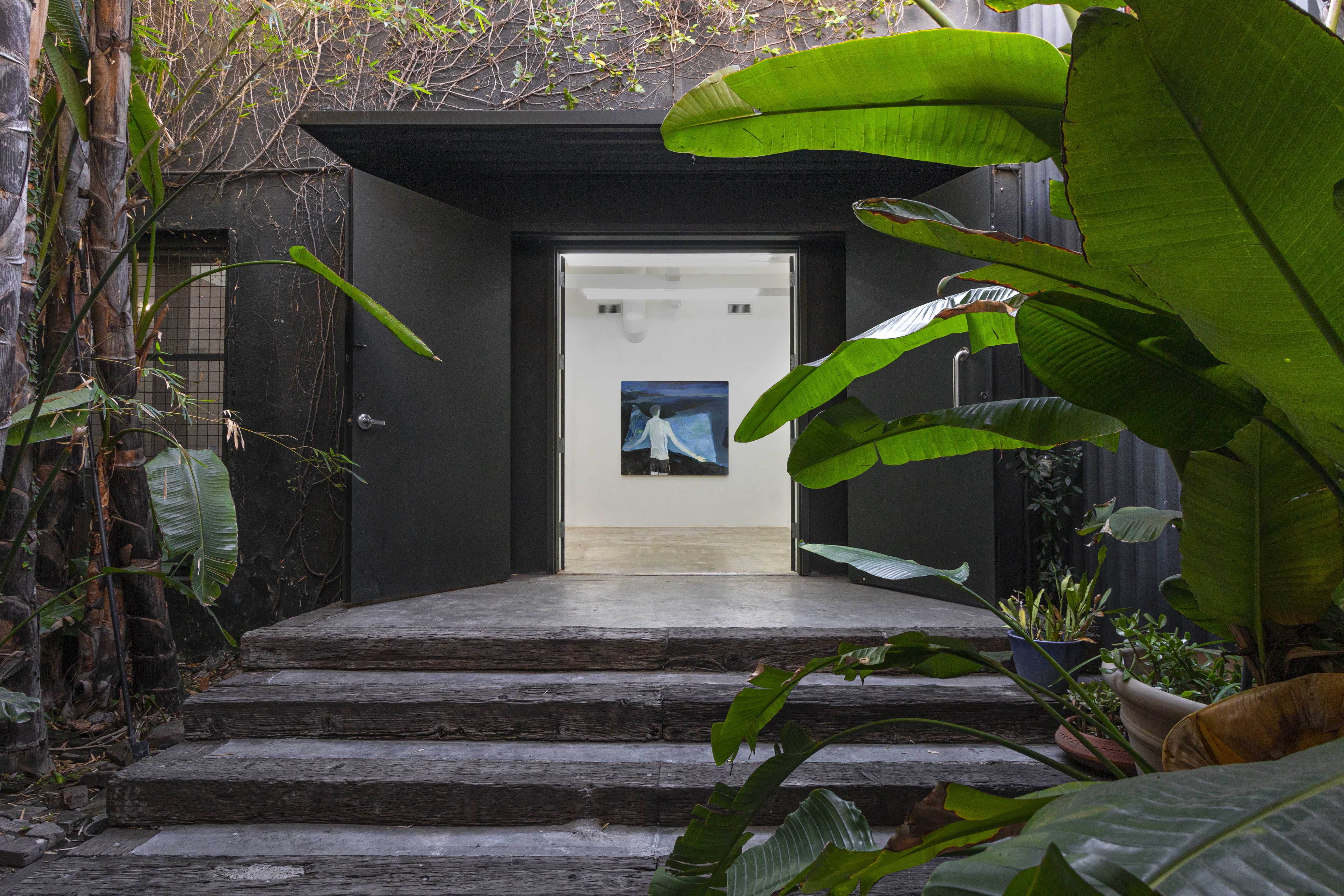 View from the outside of a gallery in which a painting hangs on the wall. The gallery is surrounded by trees and plants.